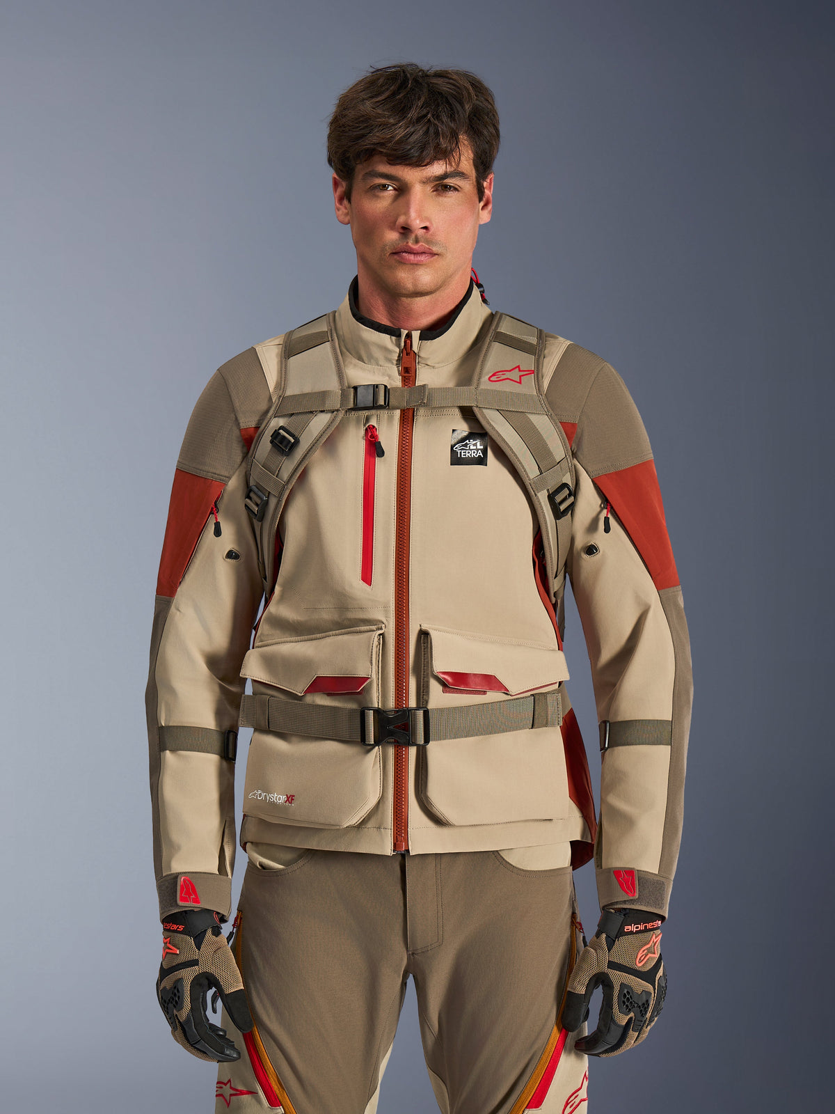 Adventure-rijder draagt de Alpinestars AT-15 Rugzak in Aluminum Walnut, vooraanzicht met borst- en taillebanden met magnetische sluiting, gecombineerd met een beige en rode adventure motorjas en -broek, technische off-road uitrusting