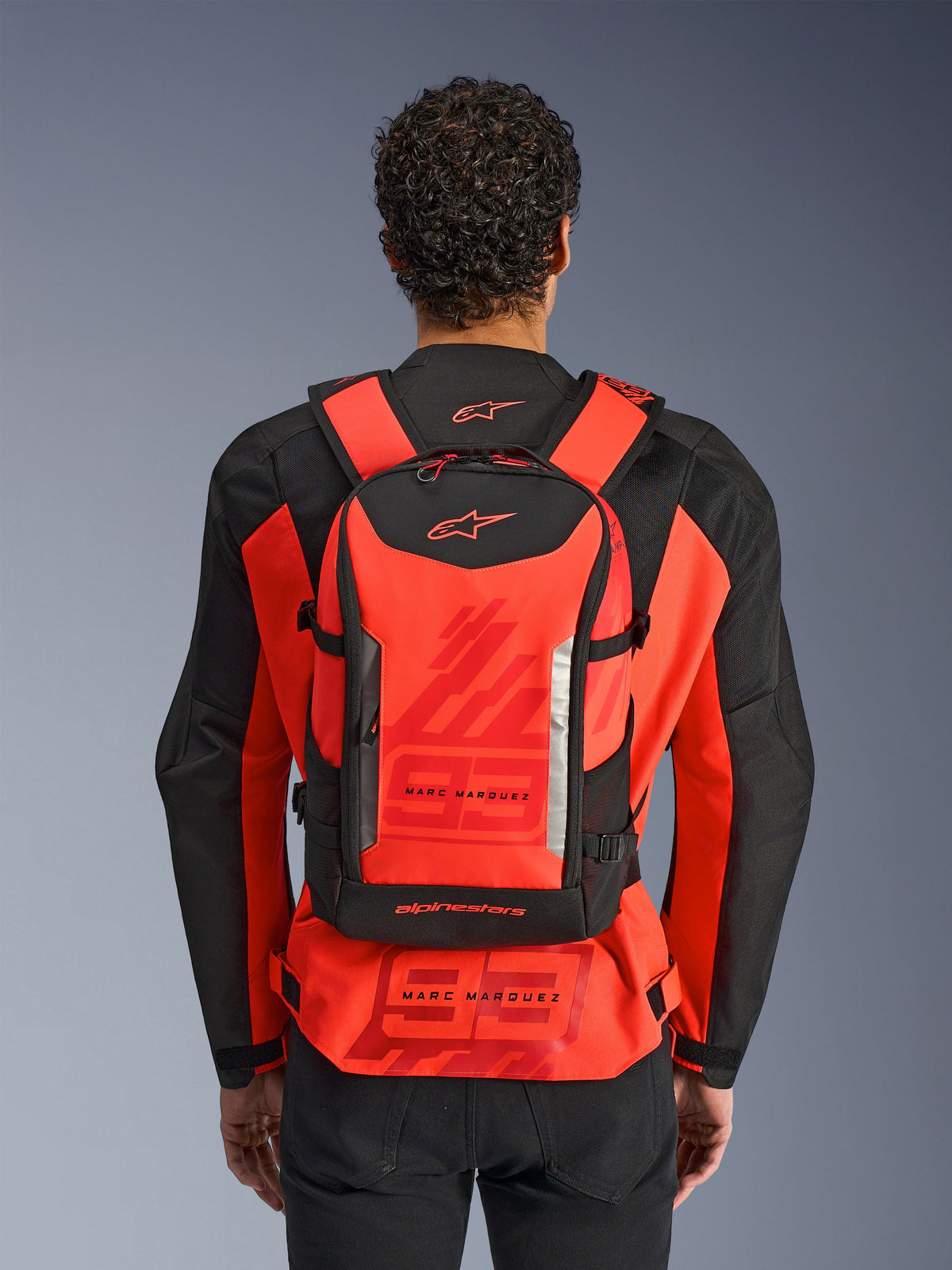 Sac à dos Alpinestars MM93 AMP3, sac à dos de moto technique, rouge fluo et noir, porté par un pilote de dos sur une veste de course MM93 rouge et noire assortie, avec marquages Marc Marquez et logos Alpinestars.