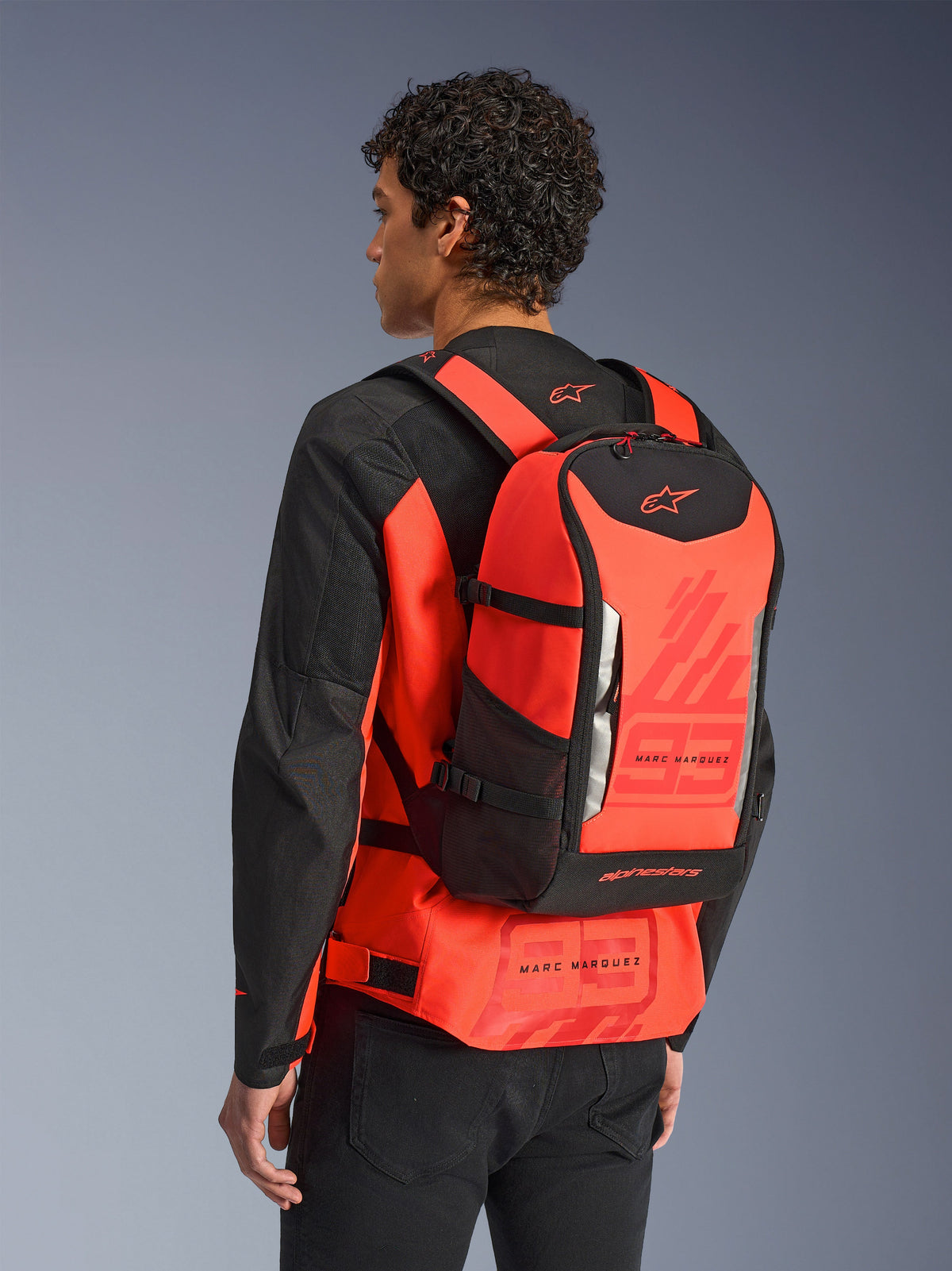 Sac à dos Alpinestars MM93 AMP3, sac à dos technique de moto, rouge fluo et noir, porté par un mannequin de dos sur une veste assortie rouge et noire, arborant les graphismes '93' de Marc Márquez, des bretelles ajustables et un design épuré d'inspiration sportive.
