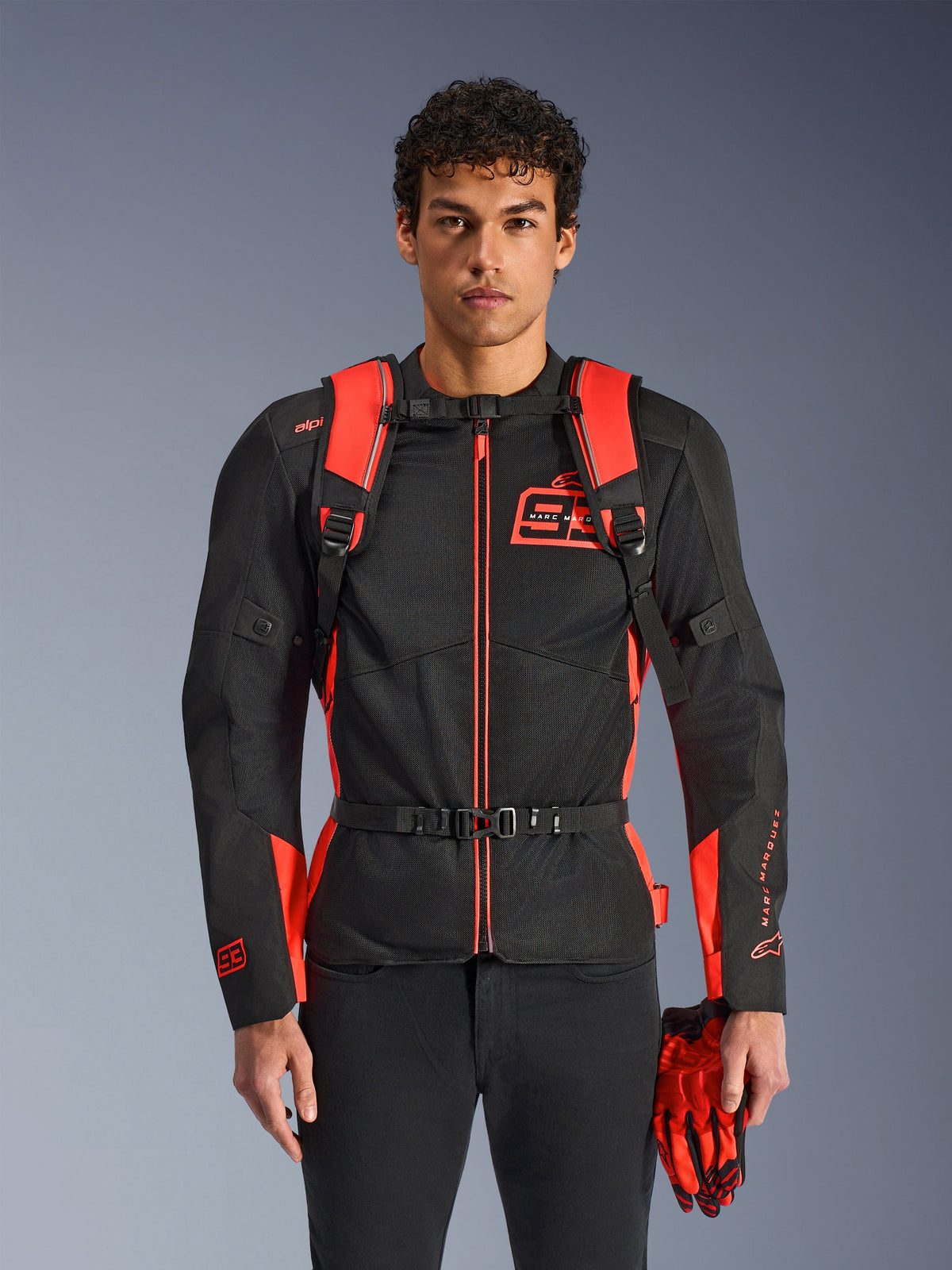 Homme portant le sac à dos Alpinestars MM93 AMP3, rouge fluo et noir, doté de bretelles réglables et d'une sangle de poitrine à boucle, assorti à une veste de moto et des gants MM93, équipement technique de la collection Marc Marquez