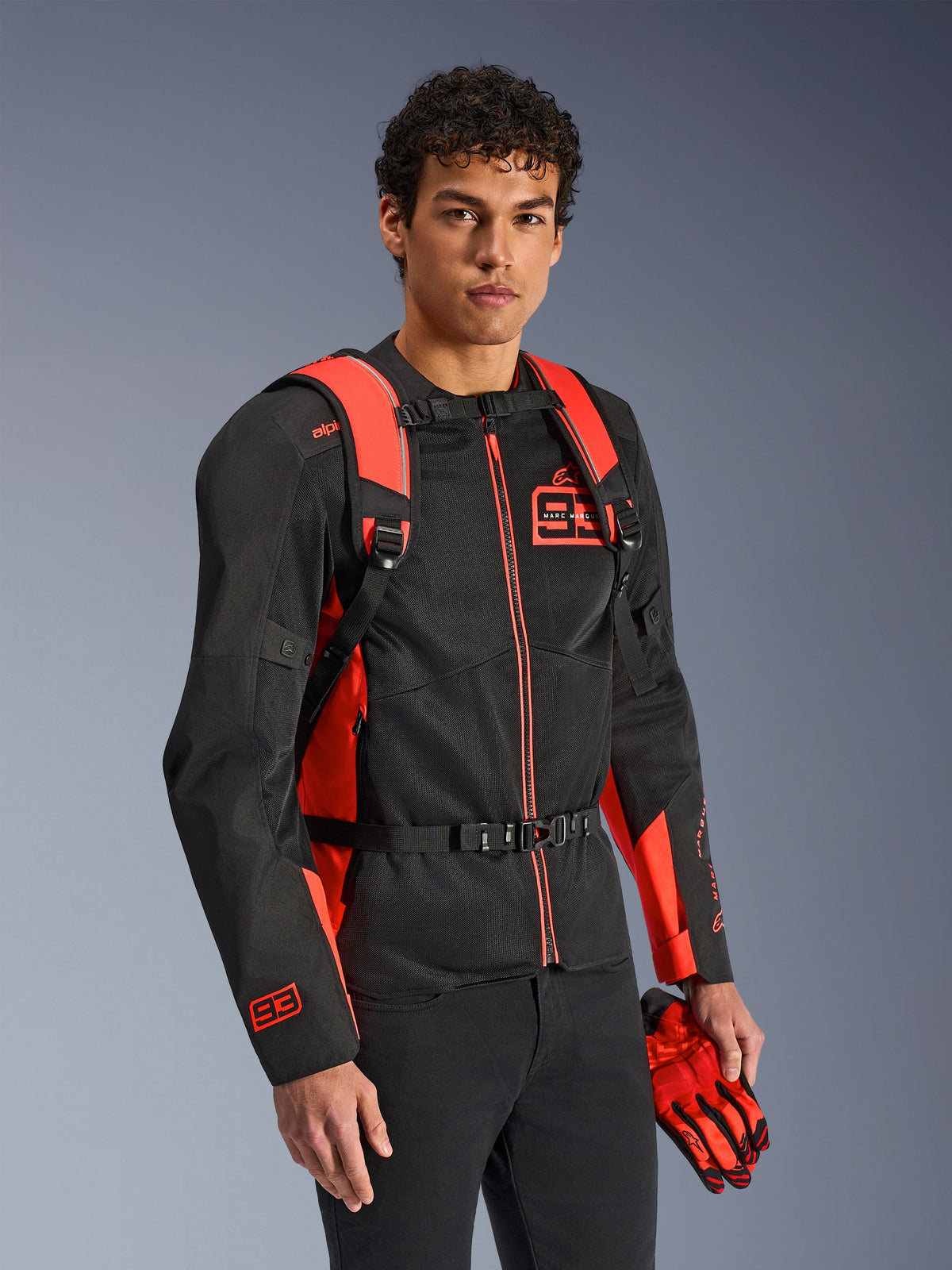 Sac à dos Alpinestars MM93 AMP3, rouge fluo et noir, porté par un modèle avec une veste de moto et des gants MM93 assortis noirs et rouges, vue de face mettant en avant les bretelles réglables et la boucle de poitrine, équipement technique inspiré de la course