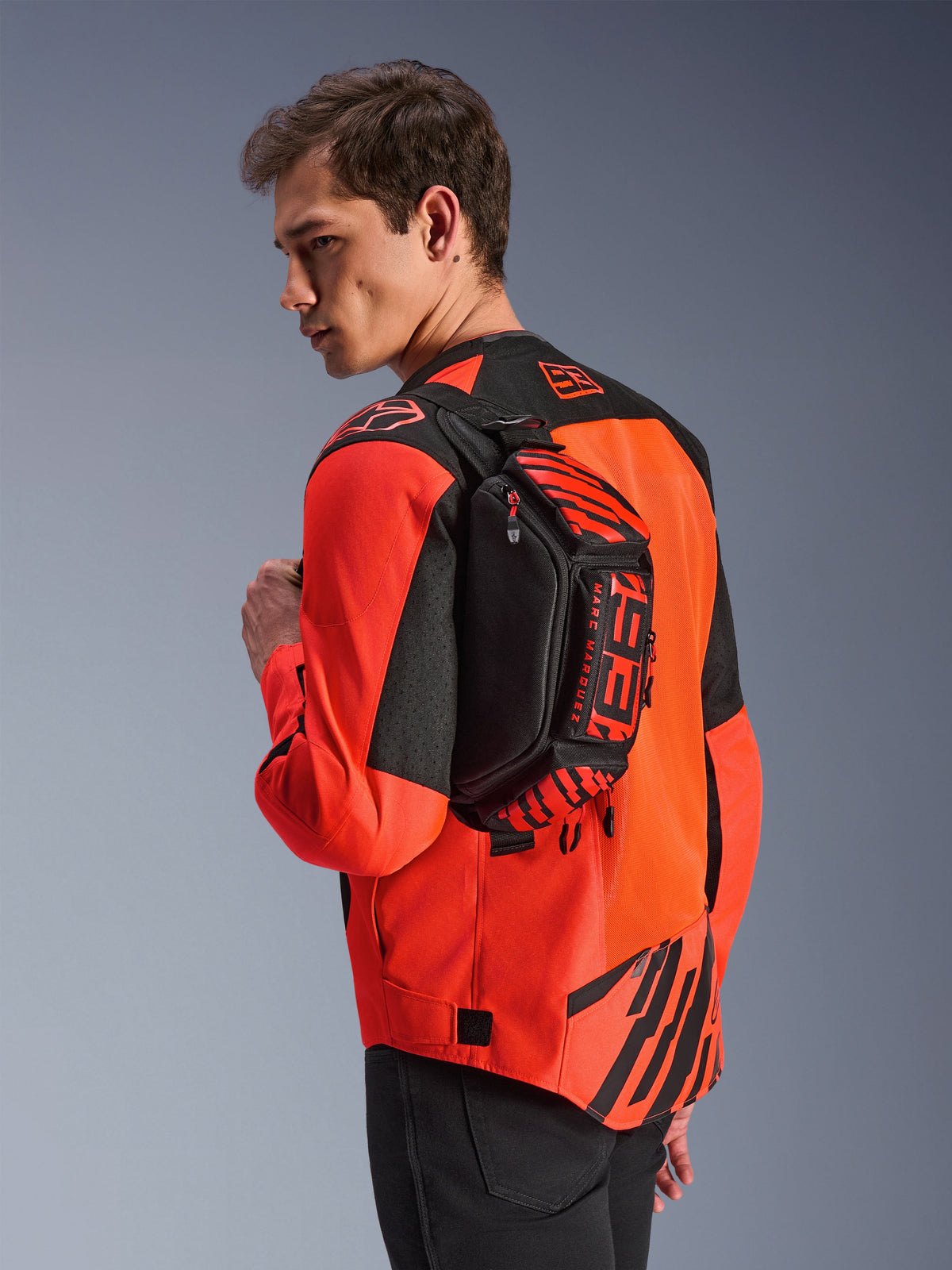 Sac banane Alpinestars MM93, noir avec rouge fluo et rouge vif, porté par un mannequin en bandoulière sur le dos par-dessus une veste de moto rouge et noire, avec logo MM93 et graphismes Marc Marquez