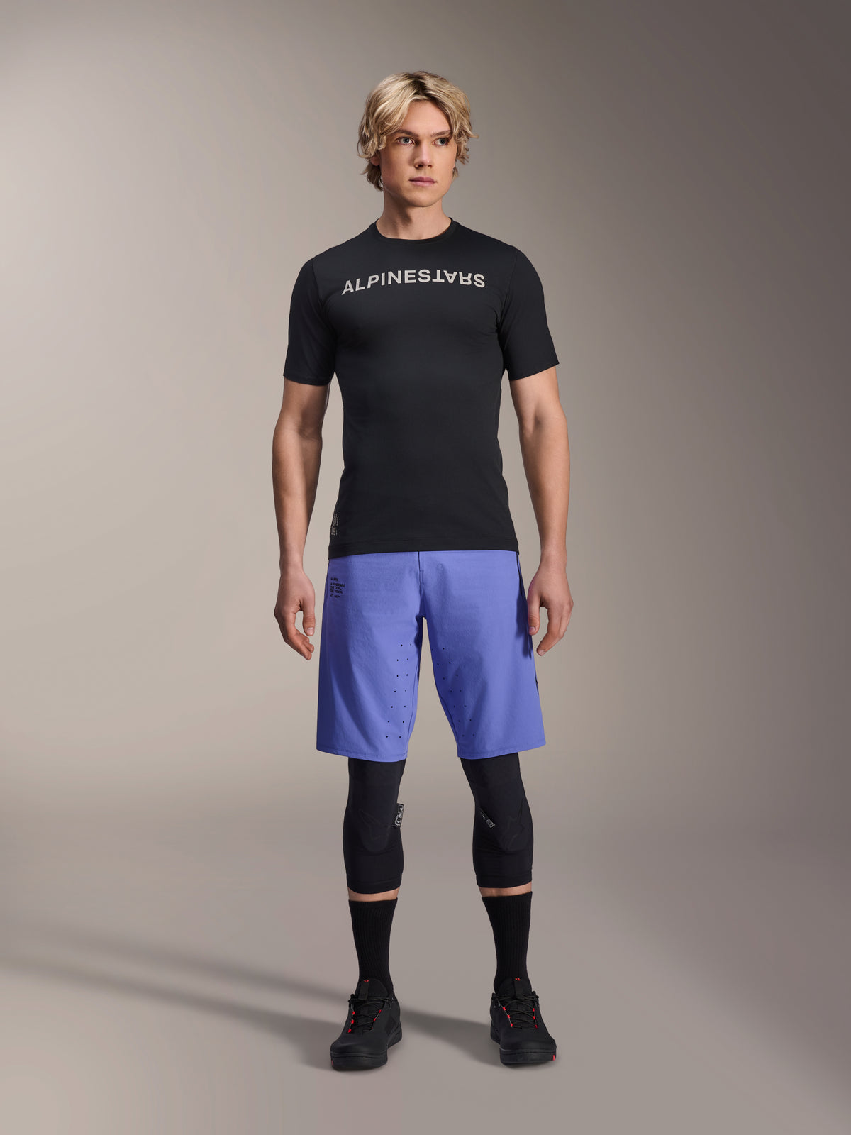Alpinestars A-Aria Switch Jersey, Short Sleeve MTB Jersey, Black with mirrored white Alpinestars logo, worn by a male model with blue cycling shorts and black knee pads, breathable performance apparel for trail riding