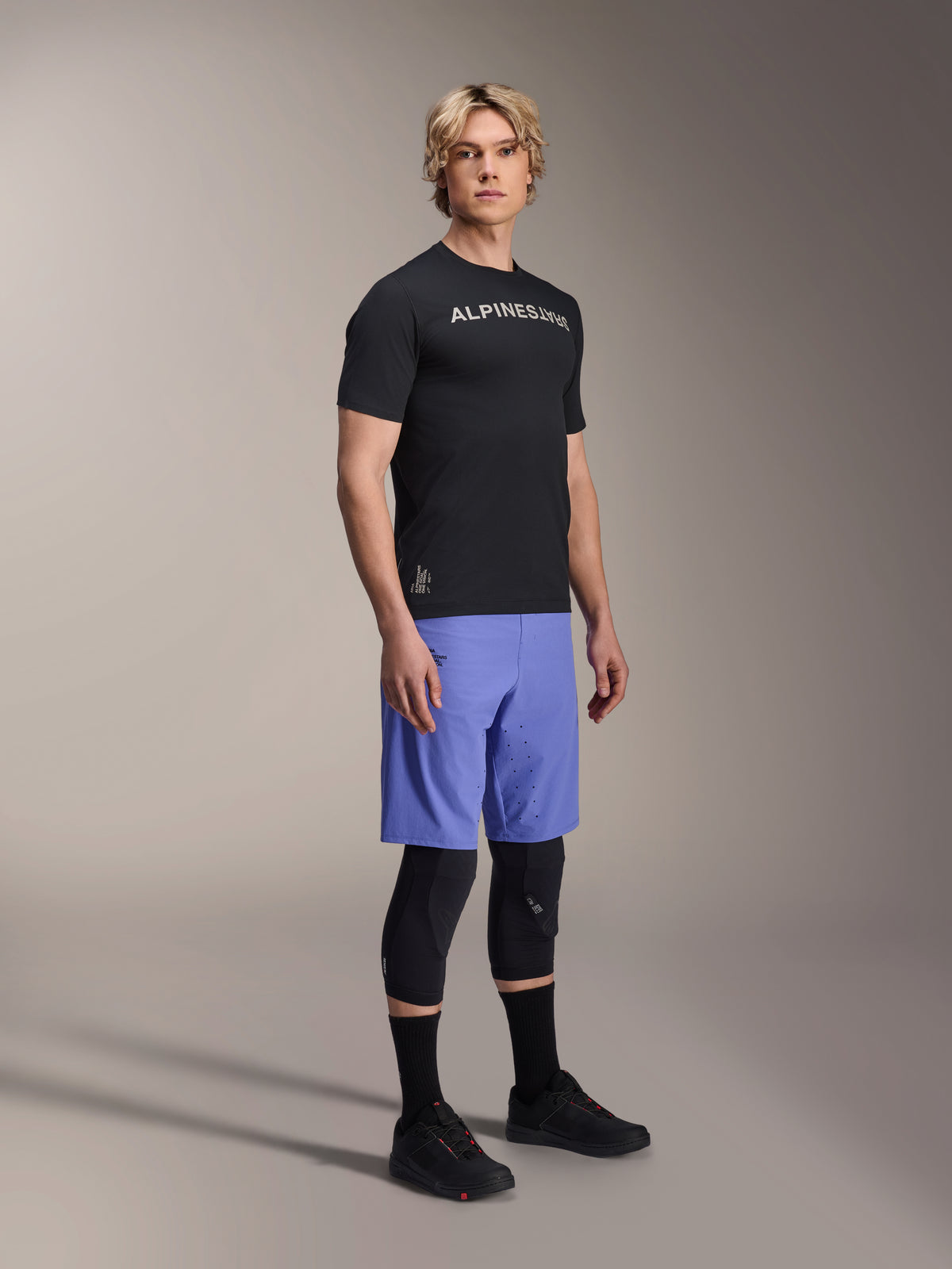 Mannequin homme portant des vêtements de VTT Alpinestars, incluant le maillot A-Aria Switch SS noir et le short A-Aria Lite bleu Baja, avec des genouillères noires, des chaussettes noires et des chaussures de VTT noires, photo de studio en pied.