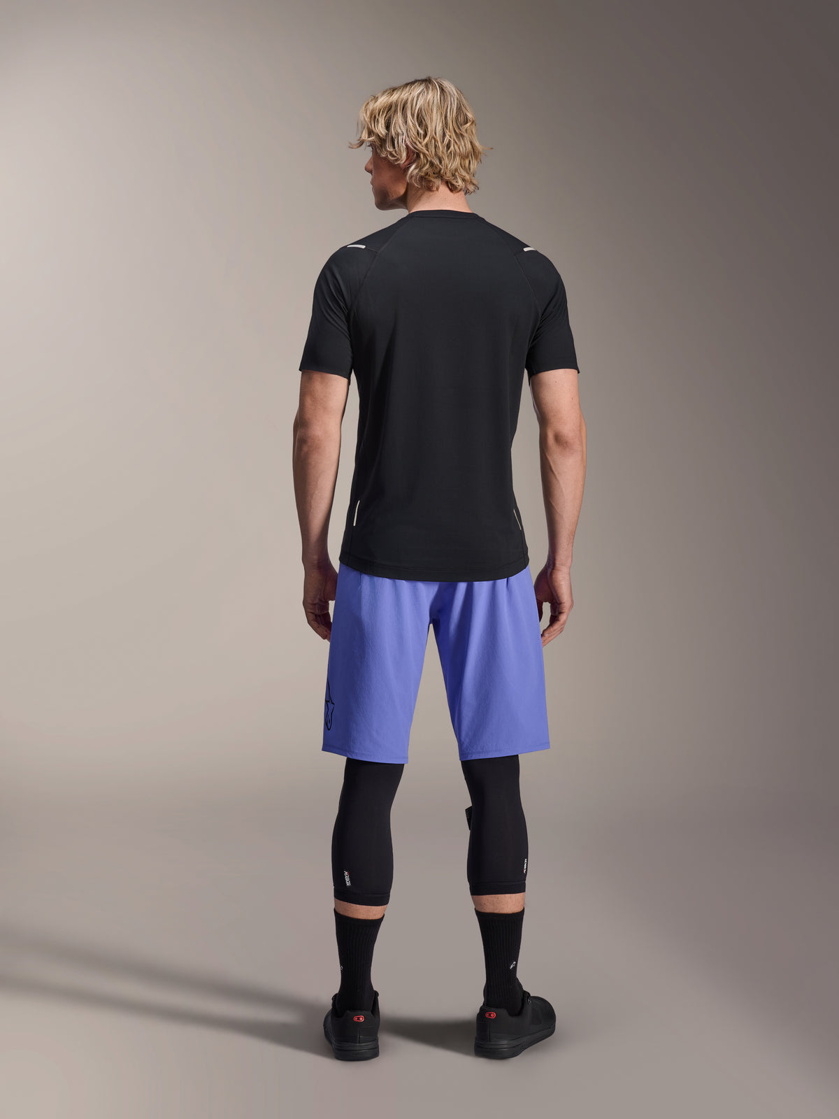 Cycliste homme portant le short Alpinestars A-Aria Lite, short de VTT, Baja Blue, vue de dos, avec maillot noir à manches courtes, genouillères noires et chaussures de cyclisme noires, équipement de VTT haute performance