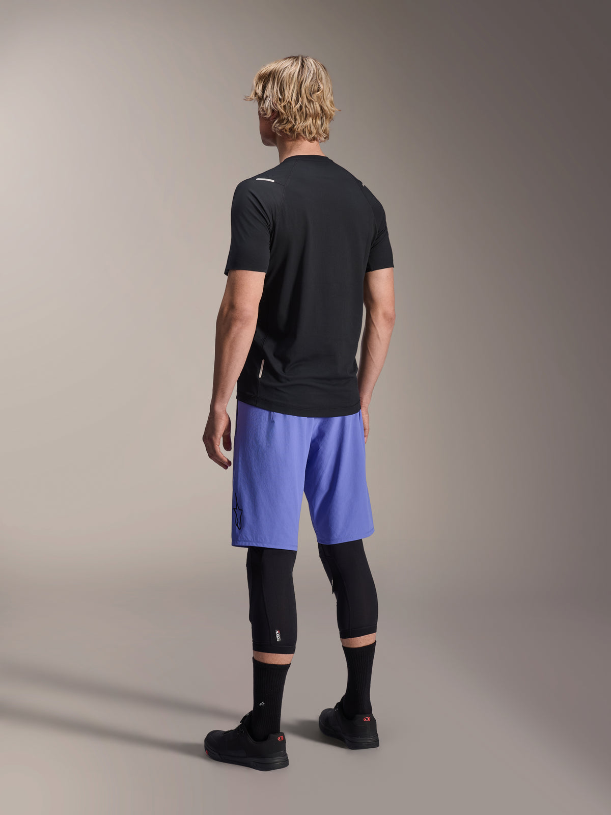 Mannequin homme de dos portant le short Alpinestars A-Aria Lite en bleu Baja, associé à un maillot noir à manches courtes et des protections de genoux noires, tenue de cyclisme VTT professionnelle pour le trail.