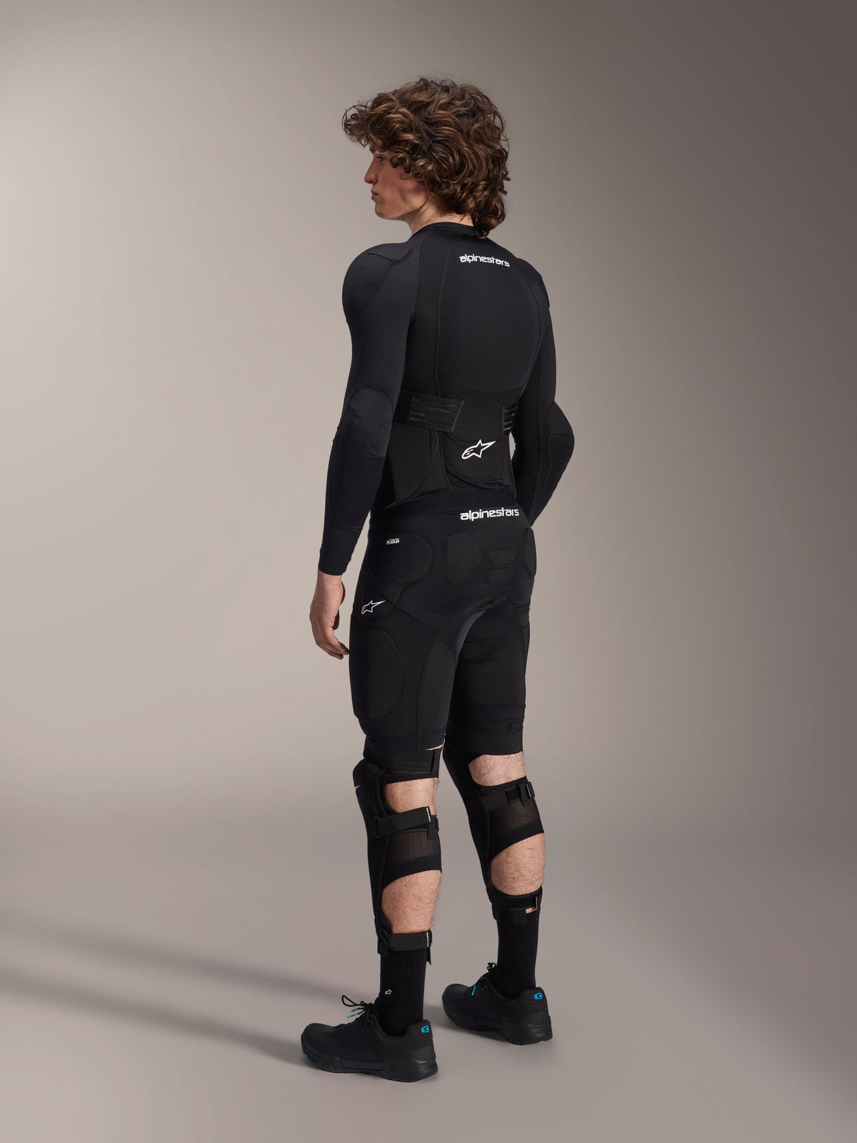 Model draagt Alpinestars A-Impact Plasma Pro knie- en scheenbeschermers vanaf de achterkant gezien, zwart en wit, toont de drievoudige riemsluiting en geventileerde mesh-panelen, gedragen met een volledige set zwarte mountainbike beschermende uitrusting inclusief een basislaag met lange mouwen en een gevoerde short