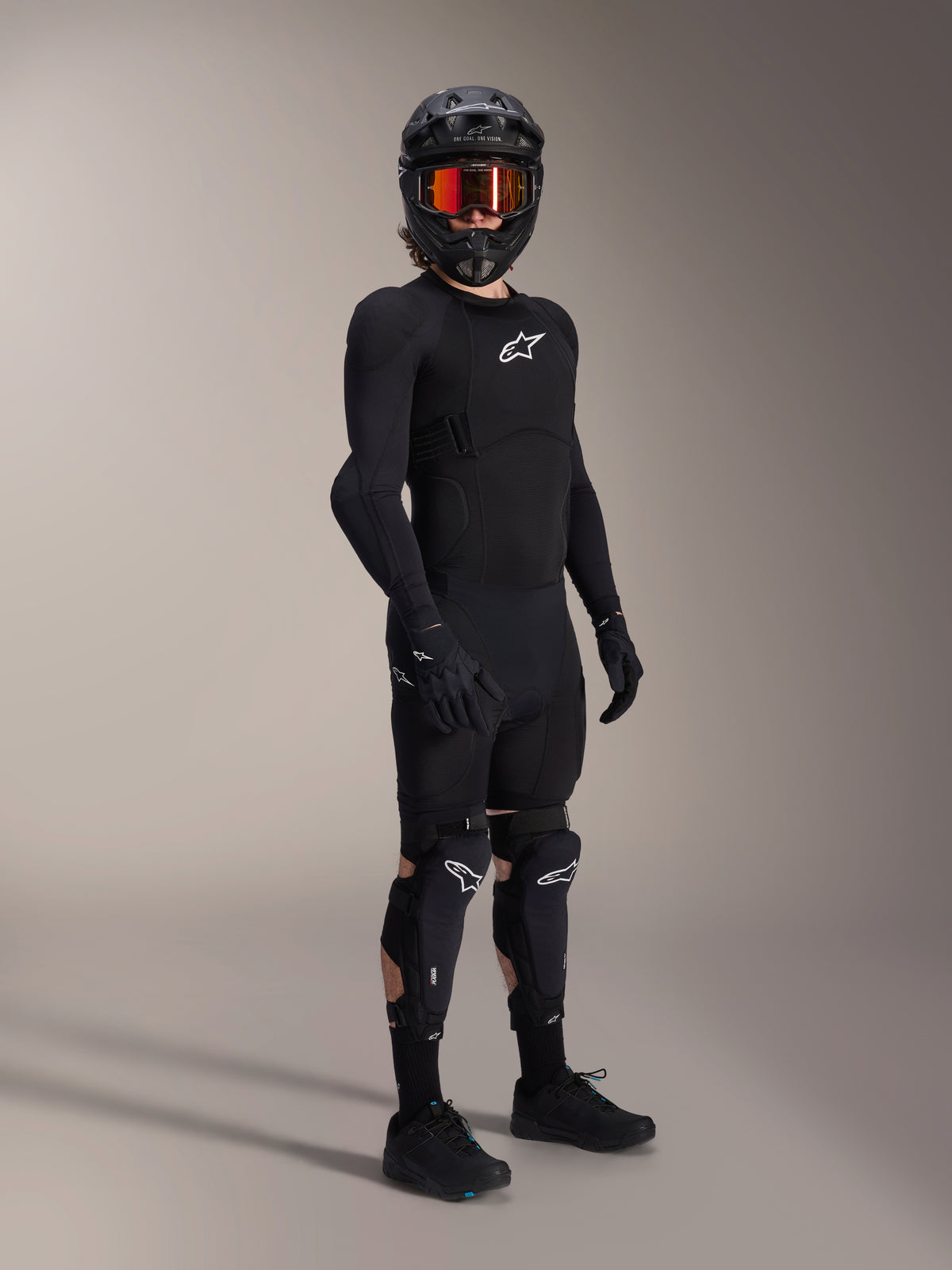 Alpinestars A-Impact Plasma Pro Knie-Schienbein-Protektoren, Schwarz und Weiß, getragen von einem Fahrer mit kompletter Mountainbike-Schutzausrüstung, inklusive schwarzem langärmeligem Protektoren-Oberteil, schwarzen Handschuhen und Fullface-Helm mit orangefarbener Goggle.