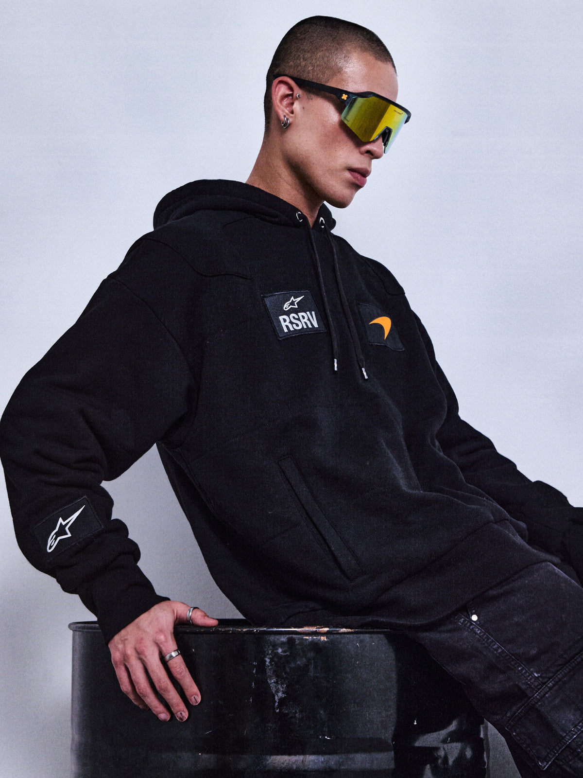 Alpinestars RSRV Rival Hoodie, sweat à capuche noir unisexe avec logo RSRV et écusson A stylisé, poche kangourou, porté avec lunettes de soleil réfléchissantes jaunes, style streetwear urbain premium