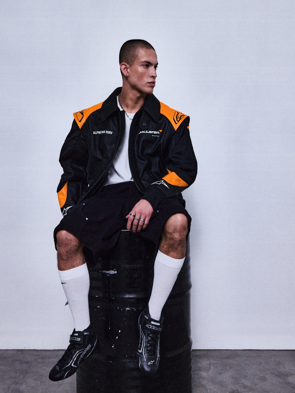 Alpinestars RSRV Track Star, blouson en cuir de moto, noir et orange, style sportif décontracté, porté avec short noir et chaussettes blanches, logos sur la poitrine