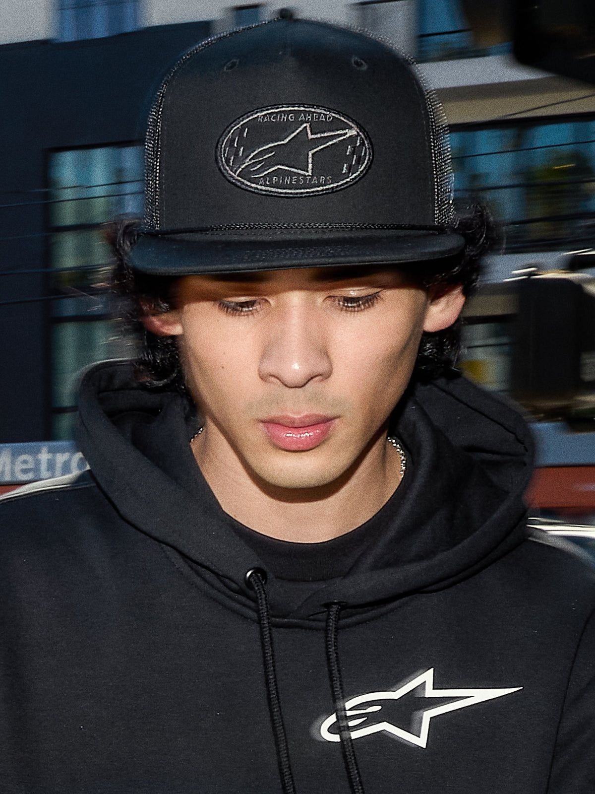 Casquette Trucker Alpinestars Contender, casquette lifestyle noire avec arrière en maille polyester, couronne structurée avec écusson brodé du logo Astars, portée par un homme avec un sweat à capuche Alpinestars noir, streetwear décontracté inspiré du motorsport.