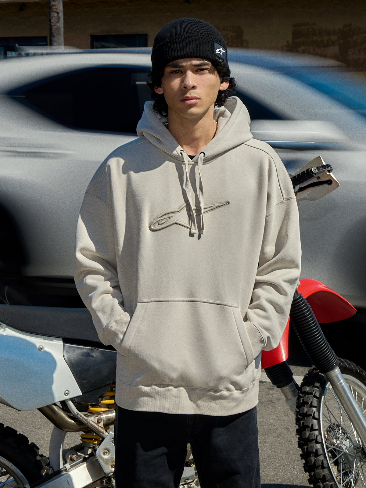 Alpinestars Emergent Oversized Hoodie en Off White, sweat à capuche en polaire de coupe ample avec logo Astars brodé en 3D, poche kangourou et capuche à cordon de serrage, porté par un homme avec un bonnet noir à côté d'une moto, streetwear casual lifestyle motorsport