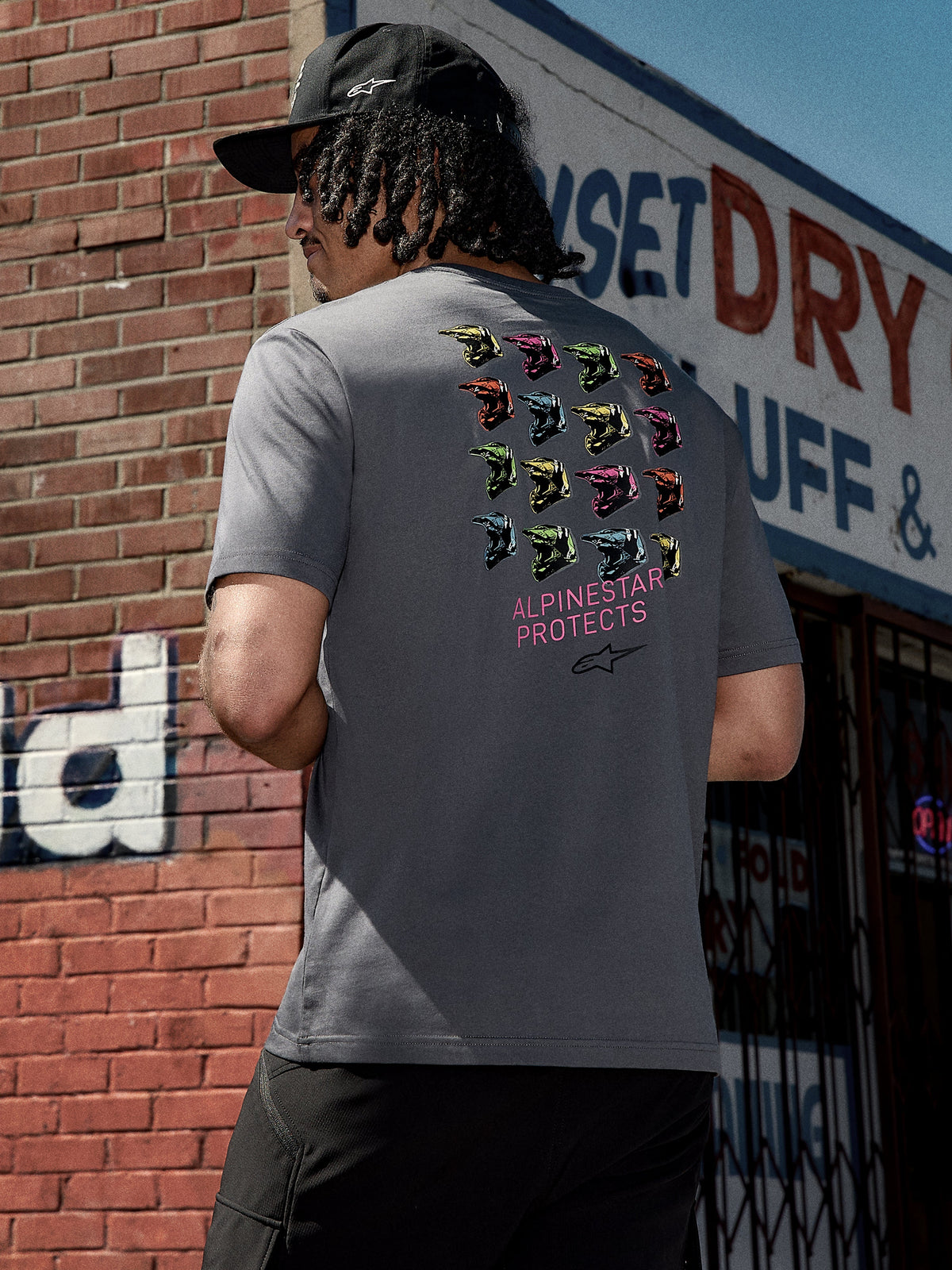 Alpinestars Pop Helmet CSF Tee, T-shirt met korte mouwen, antracietgrijs, grafische print op de achterzijde met kleurrijke M10-helmen en roze 'ALPINESTAR PROTECTS' tekst, casual motorsport streetwear