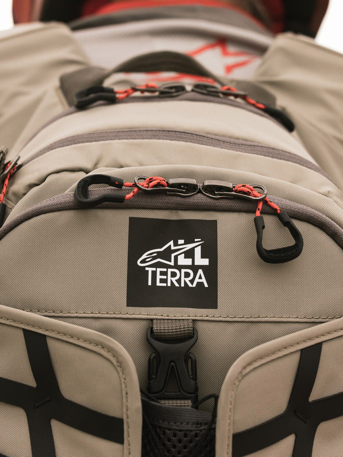 Alpinestars AT-15 Rugzak, Aluminum Walnut, close-up van de All Terra logo-patch op beige technisch textiel, voorzien van oranje trekkoorden aan de ritsen en robuuste constructiedetails