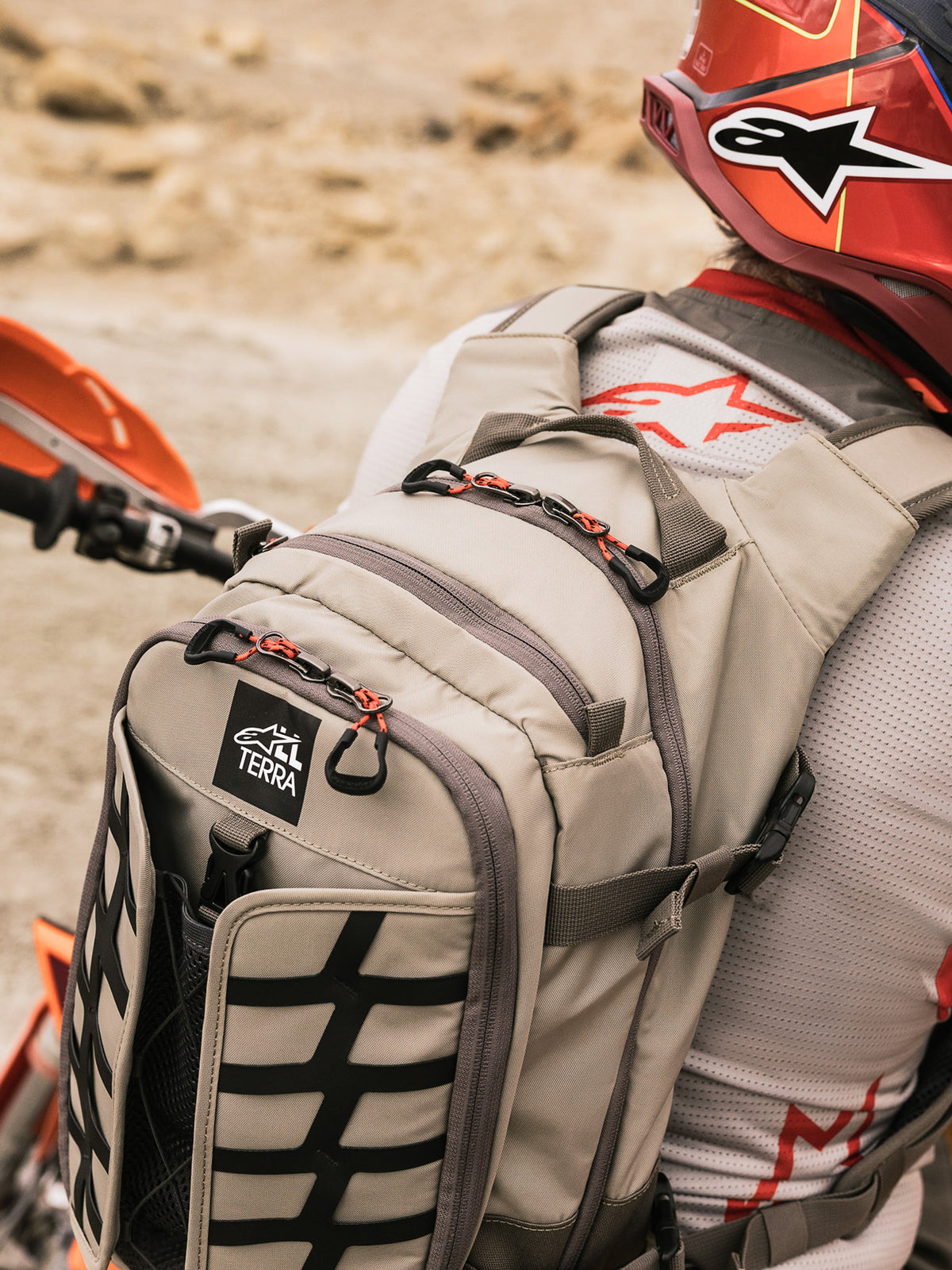 Alpinestars AT-15 Rugzak, adventure motorrugzak, Aluminum Walnut en zwart, gedragen door een rijder in een woestijnomgeving, voorzien van rode ritstrekkers, TERRA-logopatch en zwarte externe webbing, gecombineerd met een bijpassende rode helm en wit Alpinestars jersey