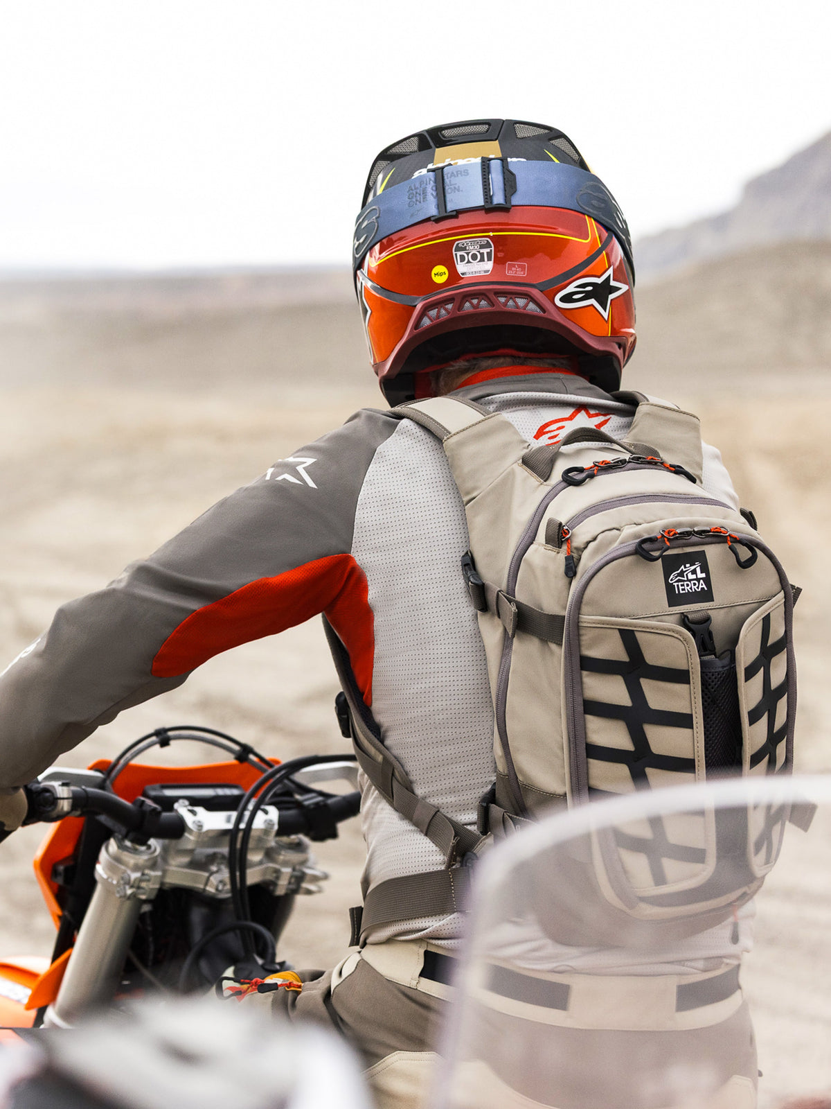 Alpinestars AT-15 rugzak in Aluminum Walnut, beige technische motorrugzak met zwarte banden en oranje ritsaccenten, 'All Terra' branding, gedragen door een adventure motorrijder in een woestijnlandschap met een rood-oranje helm en grijze motoruitrusting.