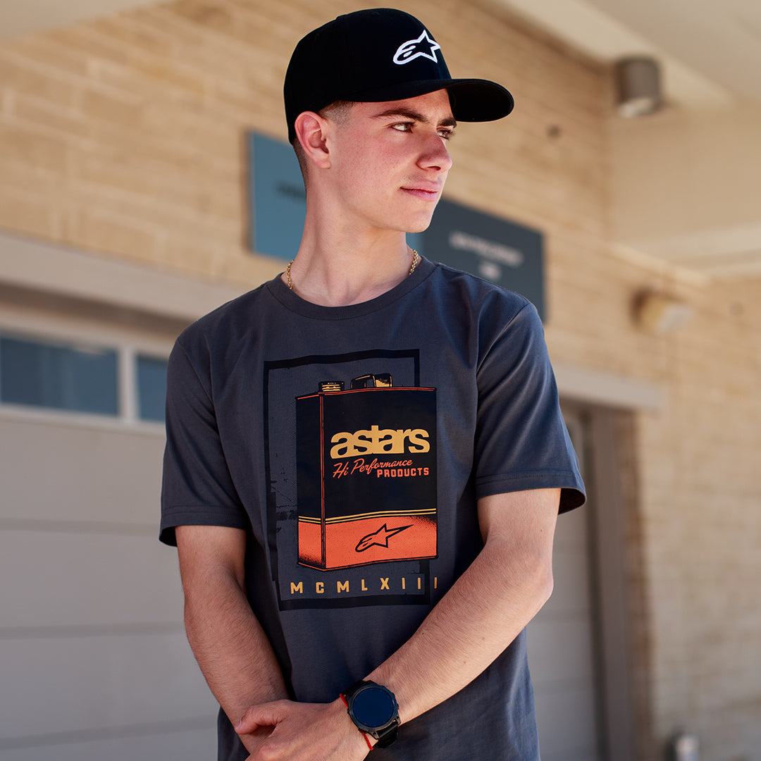 Alpinestars Galun CSF T-Shirt, Kurzarm-T-Shirt, Grau, Vintage-Benzinkanister-Grafik auf der Vorderseite mit „astars“-Branding und römischen Ziffern „MCMLXIII“, getragen von einem Mann mit schwarzer Logo-Kappe, lässiger Motorsport-Lifestyle
