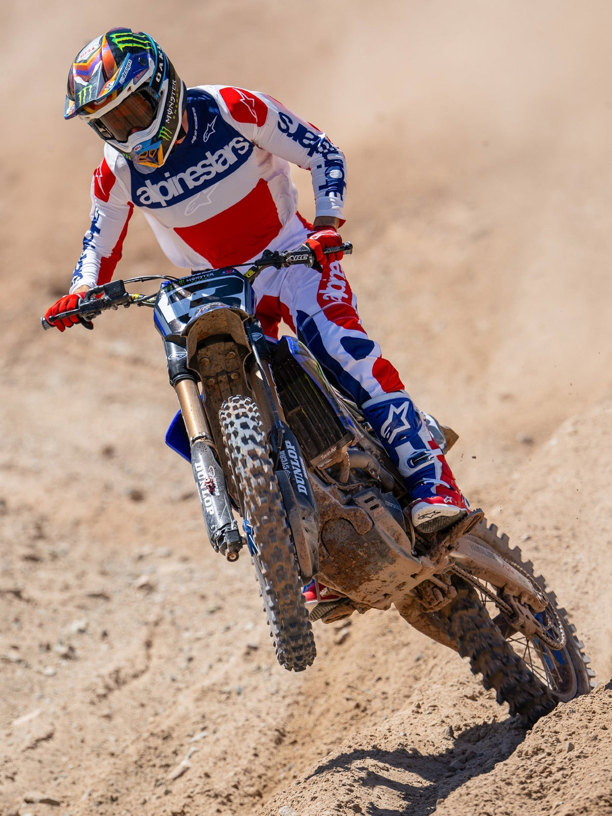 Alpinestars Limited Edition Techstar Fortitude crossbroek, motorcross broek, felrood, wit en blauw, gedragen door een rijder in actie op een zandcircuit, inclusief bijpassende jersey en laarzen, prestatiegerichte off-road race-uitrusting.