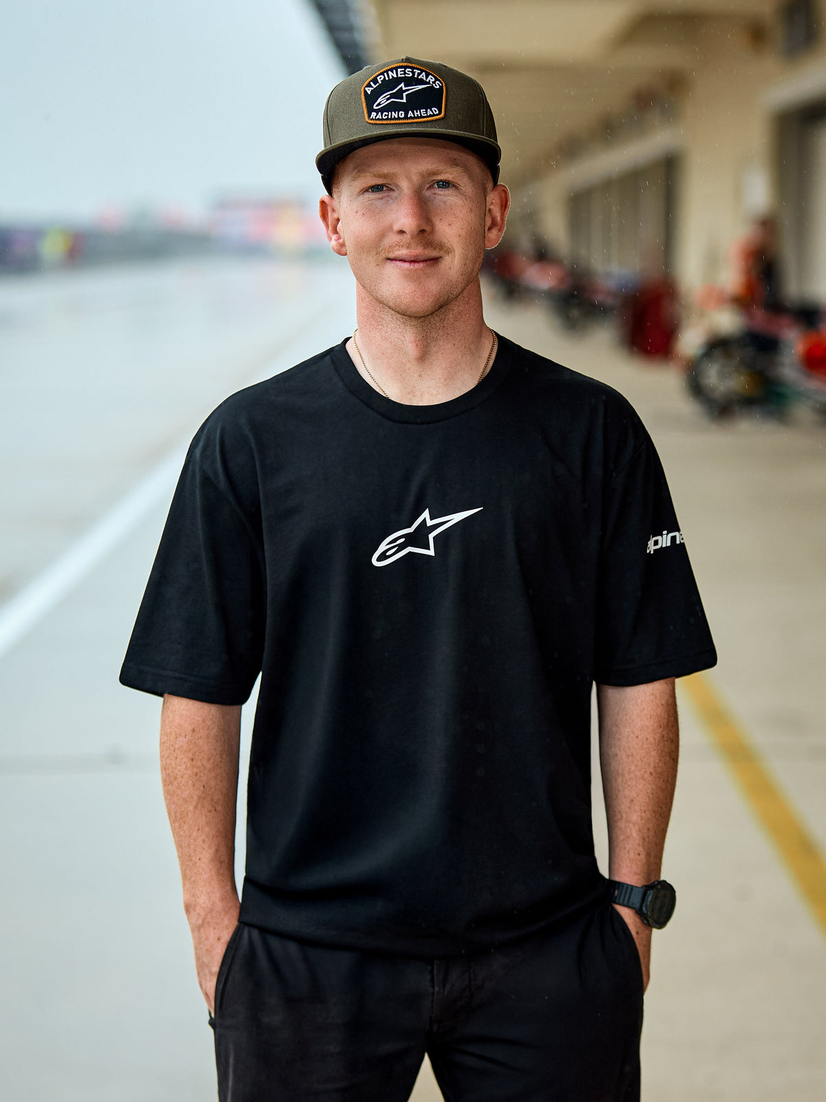 Mann im Alpinestars Frontal CSF Tee, kurzärmeliges T-Shirt, Schwarz, weißes zentrales Astars-Logo, olivgrüne Alpinestars Kappe, lässige vom Motorsport inspirierte Streetwear