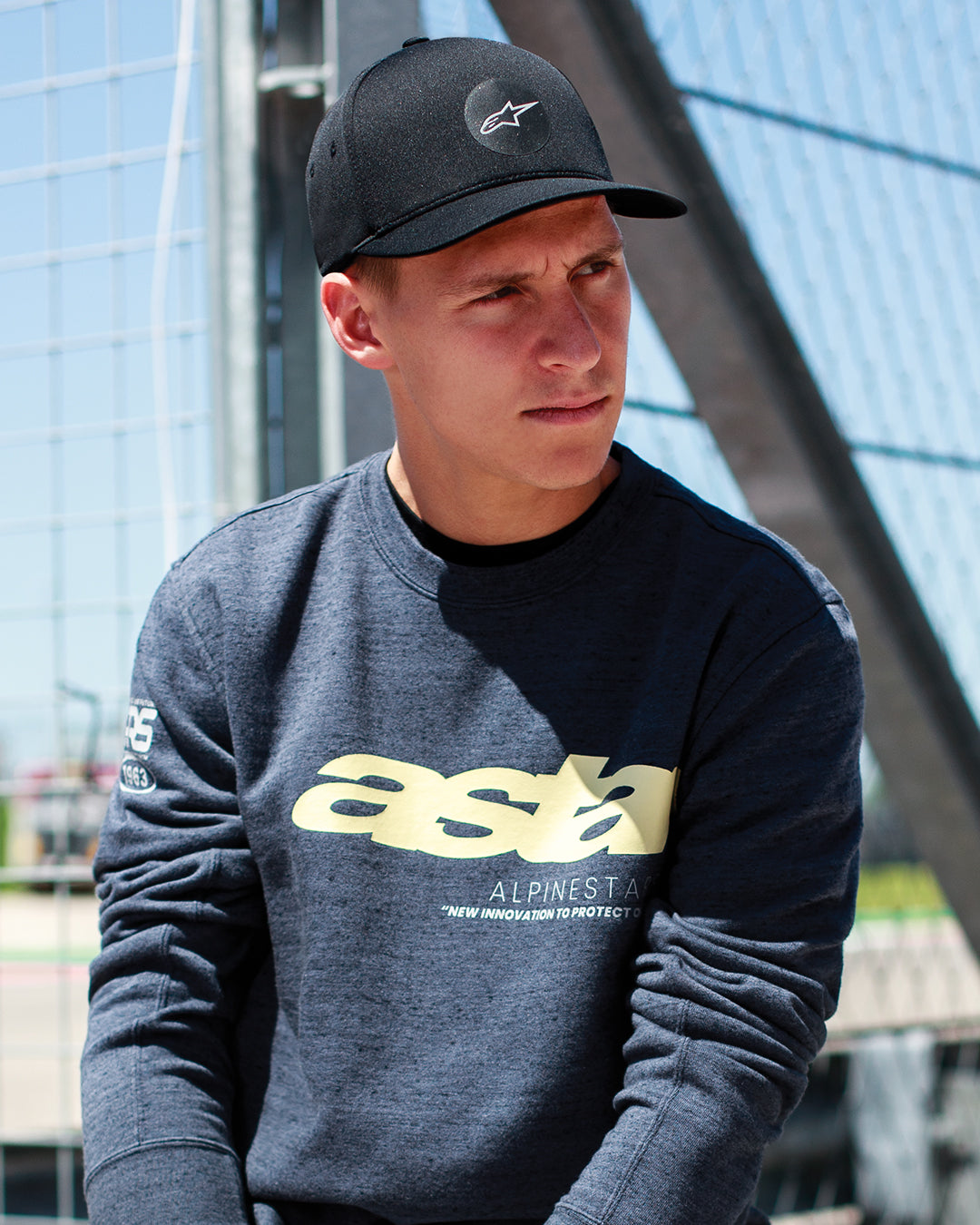 Fabio Quartararo wearing the Alpinestars Apex Hat in Black, structured crown cap with curved bill and circular star logo, paired with a dark grey heathered sweatshirt featuring yellow branding, casual motorsport-inspired lifestyle.