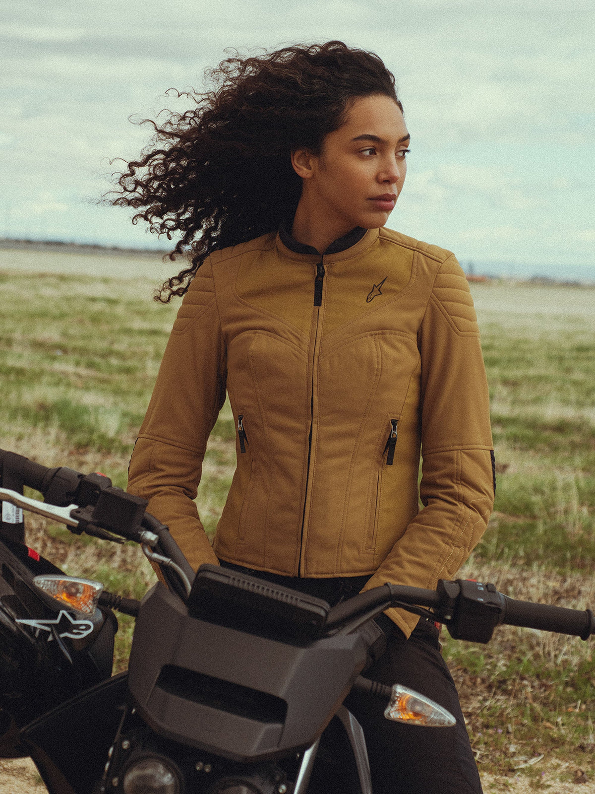 Alpinestars Isla WR Damesjas in Utility Brown Light Gold, gedragen door een vrouw op een motorfiets in een open veld, voorzien van gewatteerde schouderdetails, een slim-fit silhouet en urban contemporary styling.