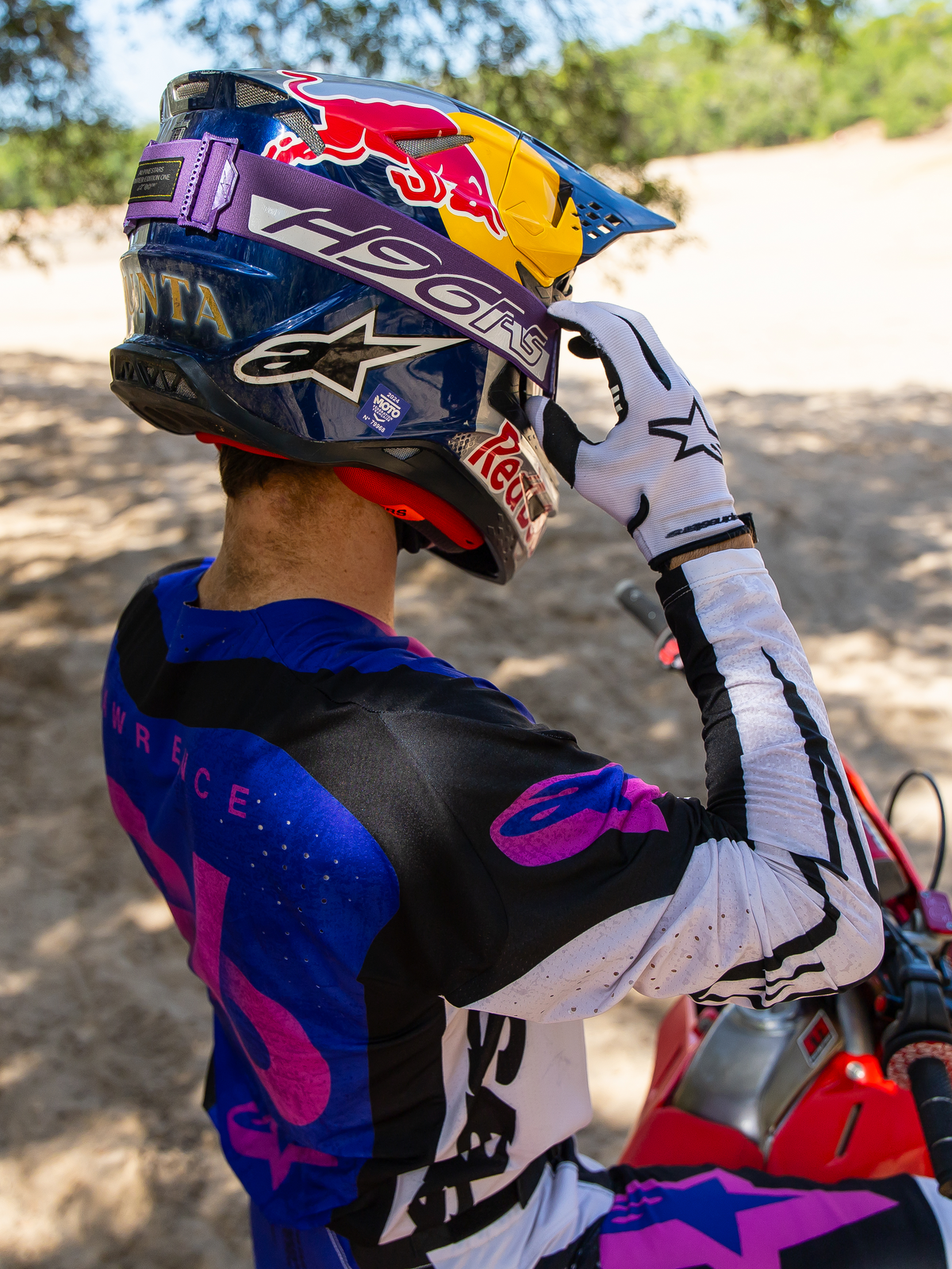 Motocross-Fahrer Hunter Lawrence mit Limited Edition HUNTA96 XXV Supertech Vision Brille, lila Band mit 'H96' und gelben Nähten, blauer Red Bull Helm, lila-weißes Trikot mit schwarzen Akzenten, weiße Handschuhe, beim Einstellen der Brille in sandiger Offroad-Umgebung