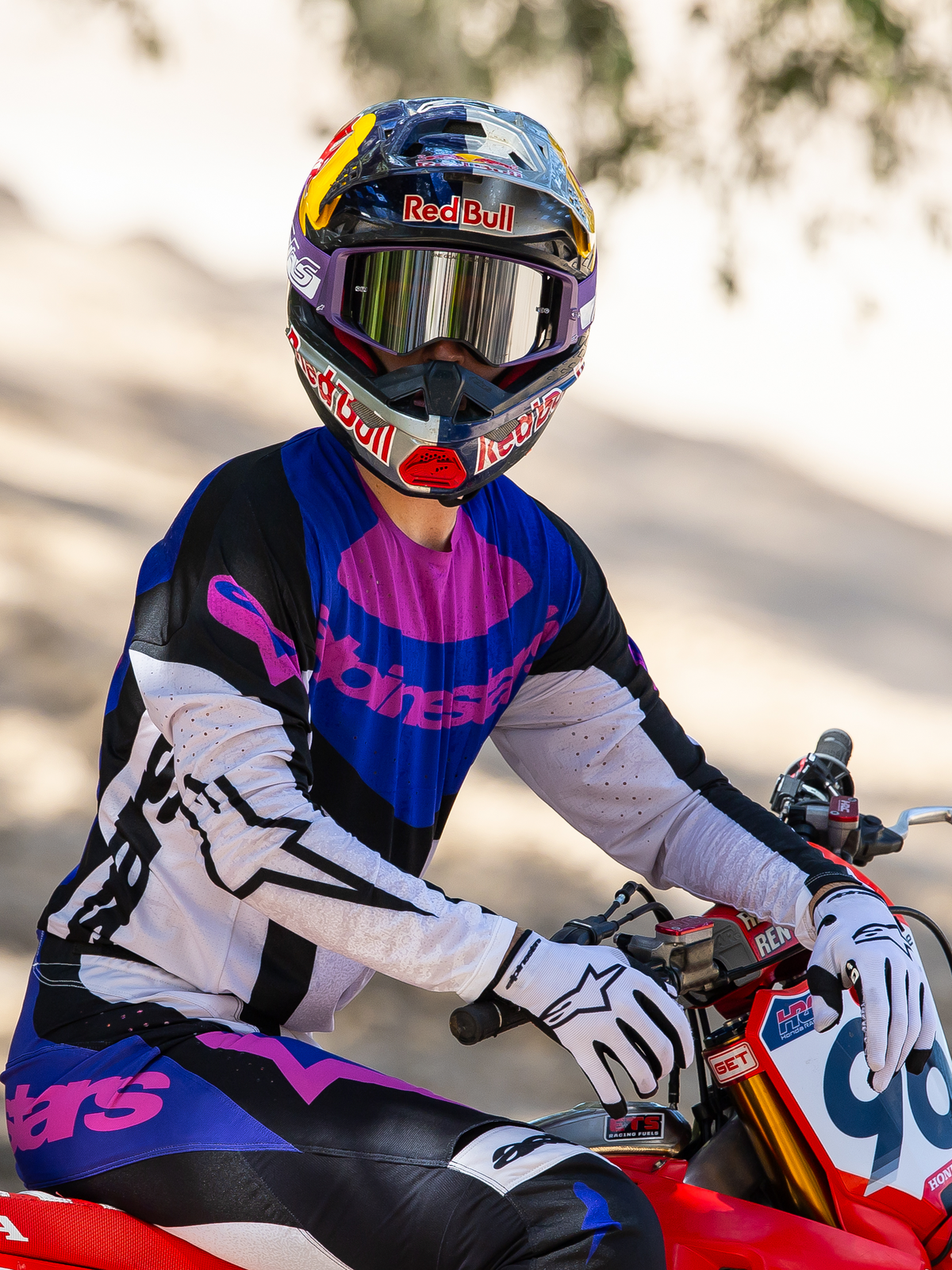 Motocross-Fahrer auf einem roten Motocross-Bike, Limited Edition HUNTA96 XXV Supertech Vision Schutzbrille in Lila und Weiß mit silbernen Spiegelgläsern, passendes lila-schwarz-weißes Alpinestars-Jersey und Hose, Red Bull-Helm, weiße Handschuhe, koordinierte professionelle Motocross-Rennausrüstung.