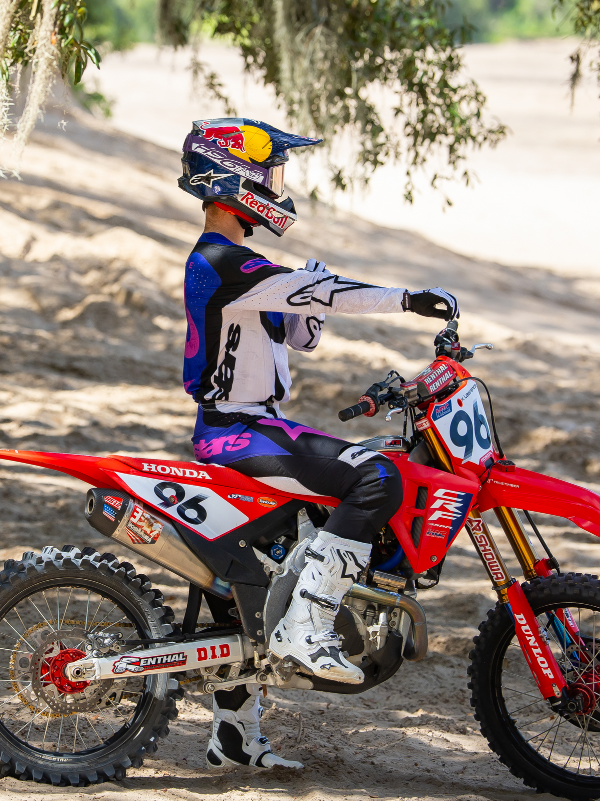 Motocross-Fahrer mit der Limited Edition HUNTA96 XXV Supertech Vision Brille in Lila, Weiß und Silber, passendes lila-weißes Renntrikot mit weißen Stiefeln, sitzend auf einer roten Honda Motocross-Maschine mit der Startnummer 96, Outdoor-Offroad-Einsatz auf einer Sandstrecke.