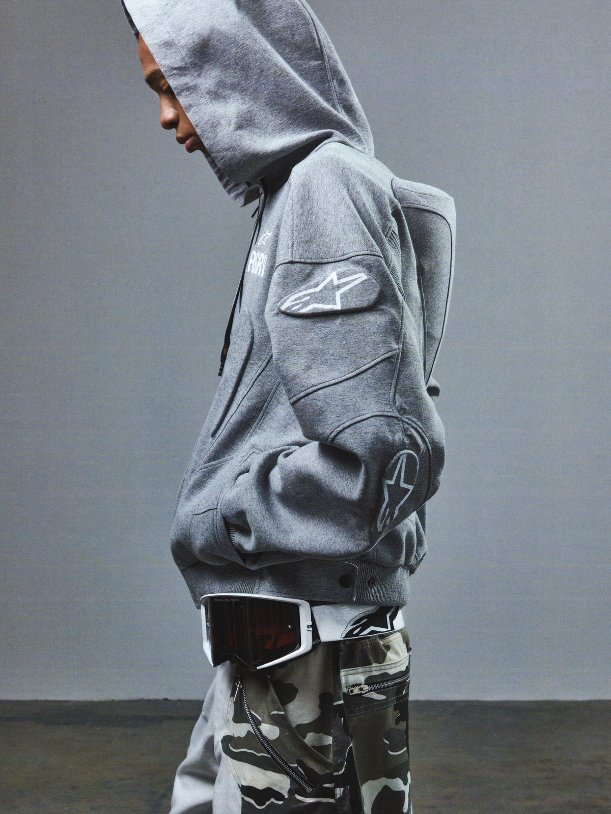 Alpinestars RSRV GP Hoodie, sweat à capuche gris avec logo étoile sur la manche, style décontracté sportif, porté avec pantalon camouflage et lunettes de protection