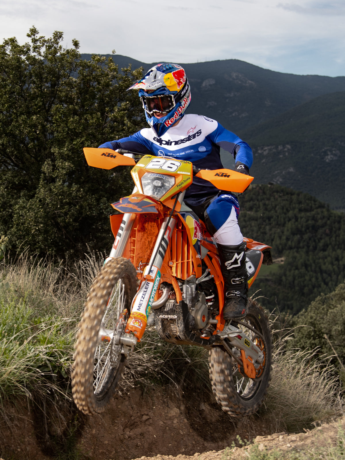 Pilote de motocross en action sur une moto KTM orange, portant l'équipement Alpinestars Pro-Dura en bleu marine, bleu brillant et blanc. Tenue tout-terrain professionnelle comprenant un maillot bleu et blanc et un pantalon assorti, avec un casque Red Bull et des bottes noires sur un sentier de montagne.