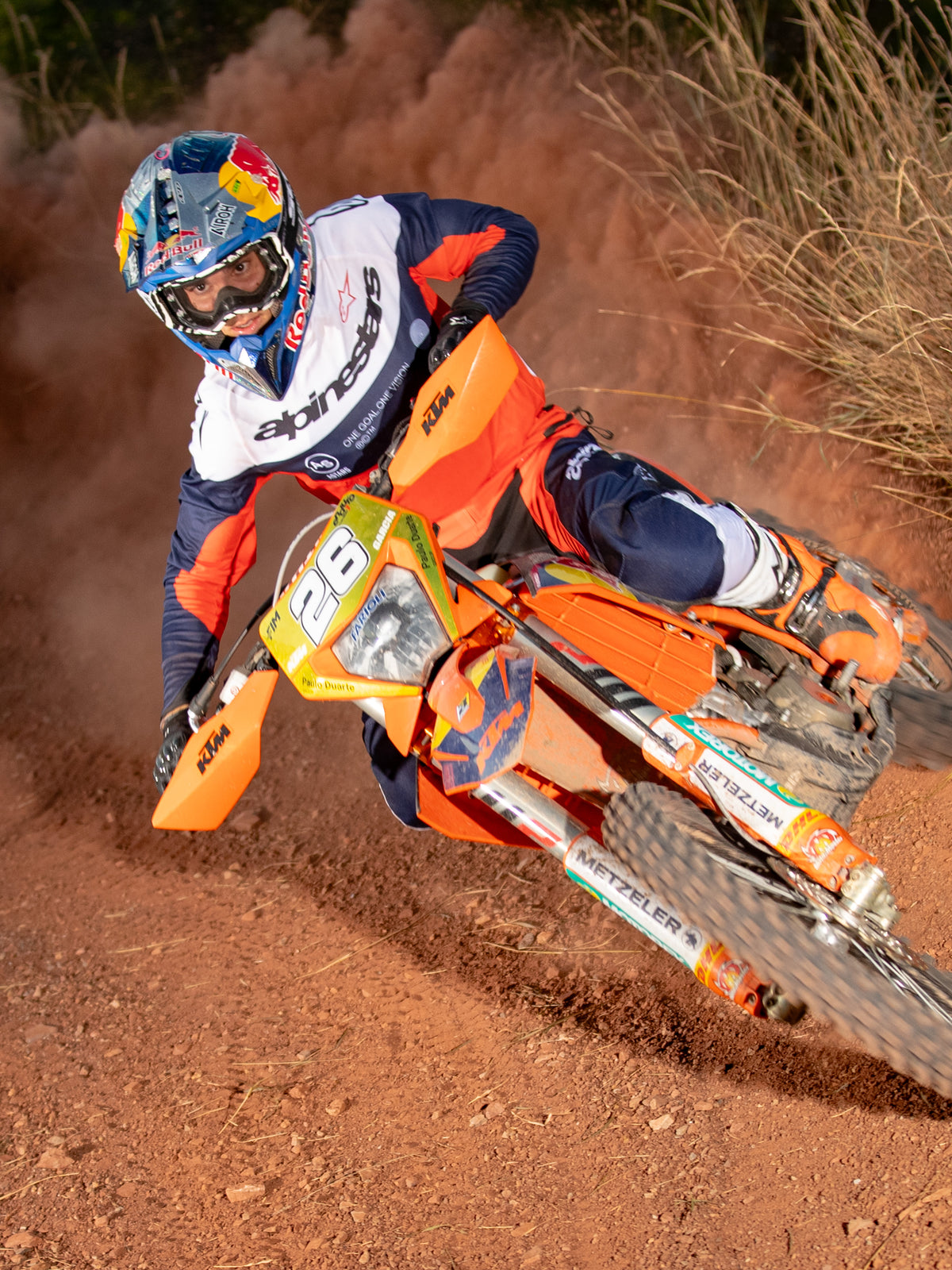 Pilote de motocross penché dans un virage sur une moto orange, portant le maillot et le pantalon Alpinestars Pro-Dura en Bleu Navy, Orange Vif et Blanc, tenue de course tout-terrain coordonnée, casque Red Bull, bottes blanches, photo d'action sur une piste de terre poussiéreuse.