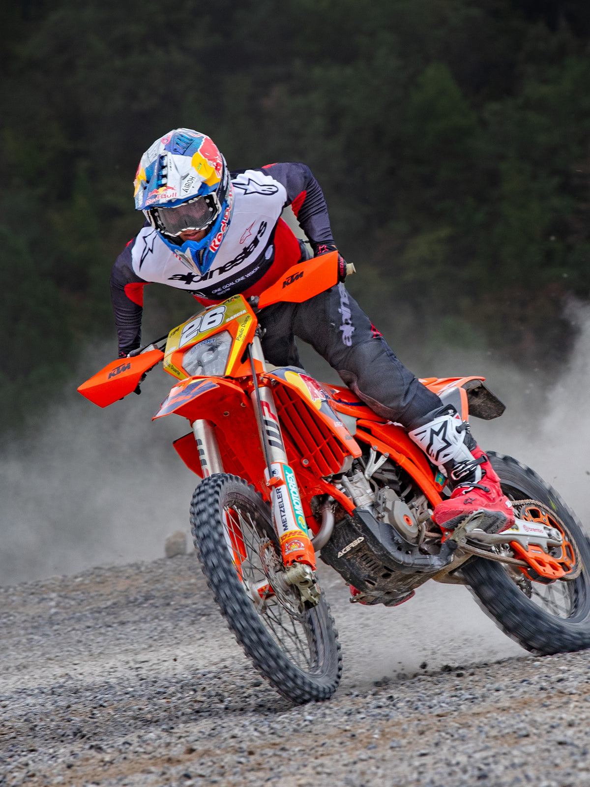 Pilote de motocross en action sur une moto KTM orange, portant l'équipement Alpinestars Pro-Dura comprenant un maillot rouge, blanc et noir et un pantalon gris foncé, des bottes rouges et blanches et un casque de course Red Bull, effectuant un virage serré sur un sentier de gravier