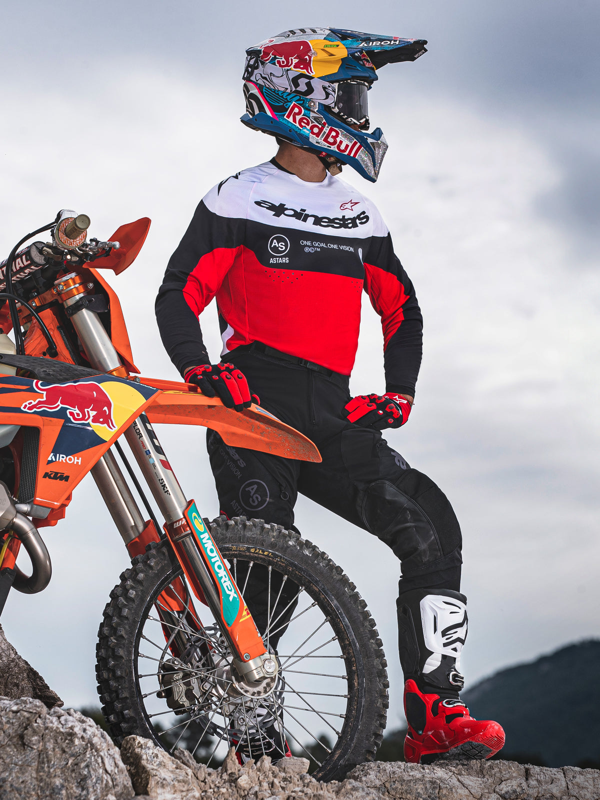 Pilote de motocross debout à côté d'une moto tout-terrain orange sur un terrain rocheux, portant l'équipement Alpinestars Pro-Dura comprenant un maillot rouge, blanc et noir et un pantalon noir, avec des gants rouges et noirs, des bottes blanches et rouges et un casque Red Bull, tenue d'enduro professionnelle complète.