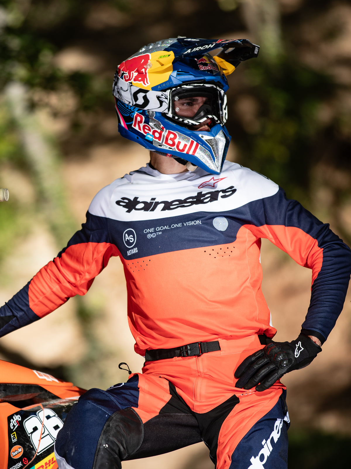Pilote de motocross portant l'équipement Alpinestars Pro-Dura, maillot bleu marine, orange vif et blanc avec logo Alpinestars central, pantalon Pro-Dura assorti, casque Red Bull et gants noirs, tenue coordonnée pour la course d'enduro professionnelle