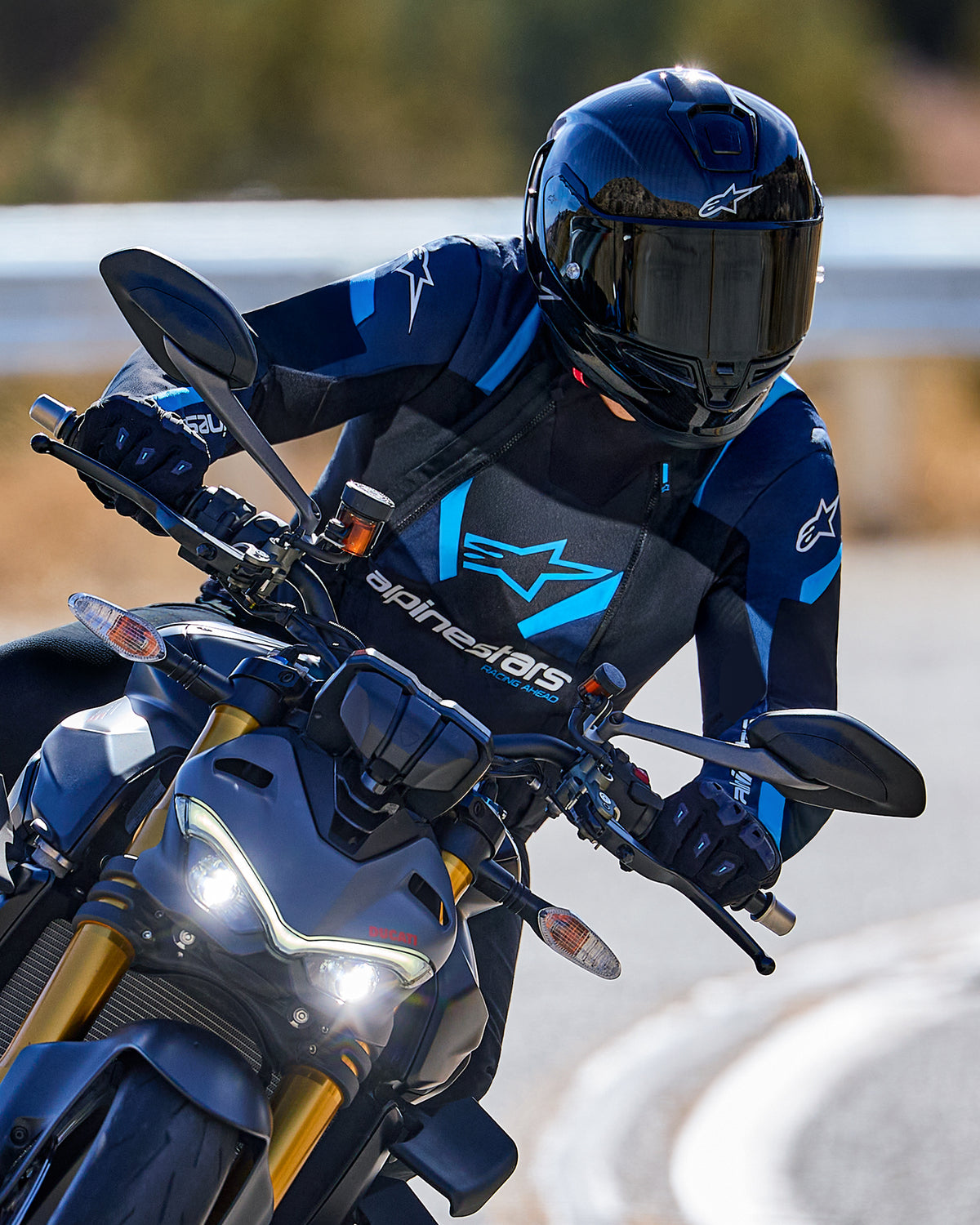 Motorcycle rider leaning into a curve wearing the Alpinestars T-Stunt Air Jacket in Black, Dark Blue, and Bright Blue, paired with a black helmet and gloves, action shot on a paved road, performance sport riding gear