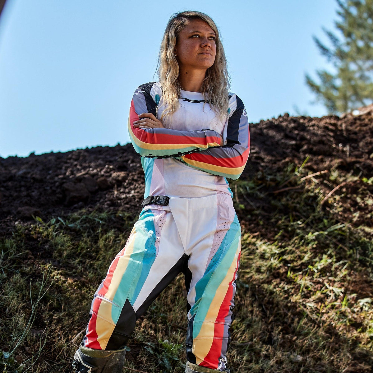 Female motocross rider wearing Alpinestars Women Stella 2024 Techstar Pants and matching jersey, Black Multicolor design with white base and vibrant blue, yellow, and red stripes, standing with arms crossed on a dirt track, professional off-road racing outfit