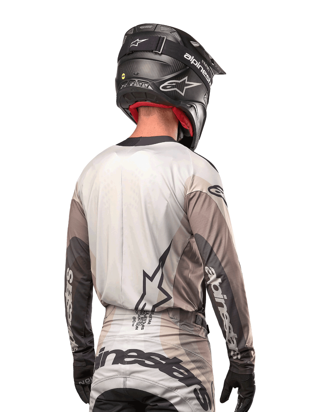 Pilote de motocross vu de dos portant le maillot Alpinestars 2024 Techstar Pneuma en Dark Sand Iron Dust Gray, associé à un casque noir à finition carbone, avec un grand logo étoile dans le dos et le marquage Alpinestars sur les manches.