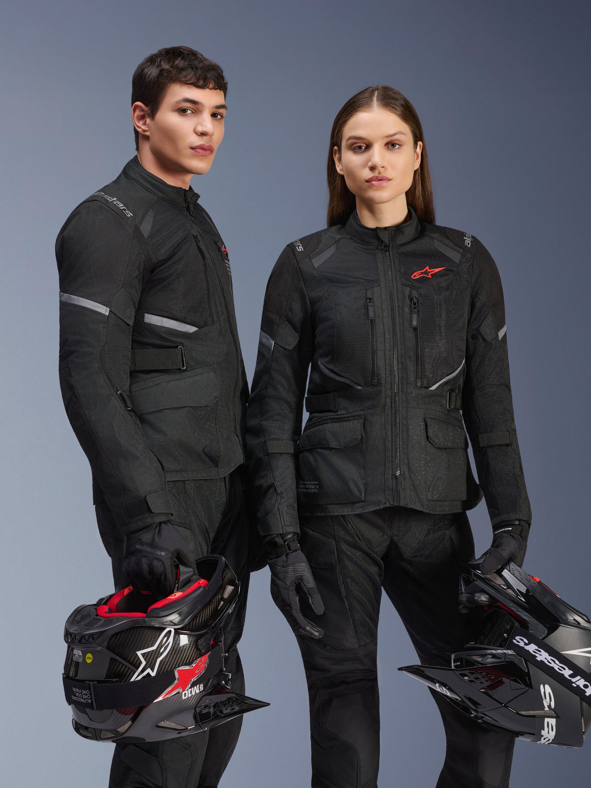 Modèles homme et femme portant des vestes Alpinestars Stella Andes Air Drystar® en noir, équipement de moto touring aventure avec logos d'étoile rouges, panneaux de ventilation en maille et construction textile technique, modèles tenant des casques d'aventure en studio