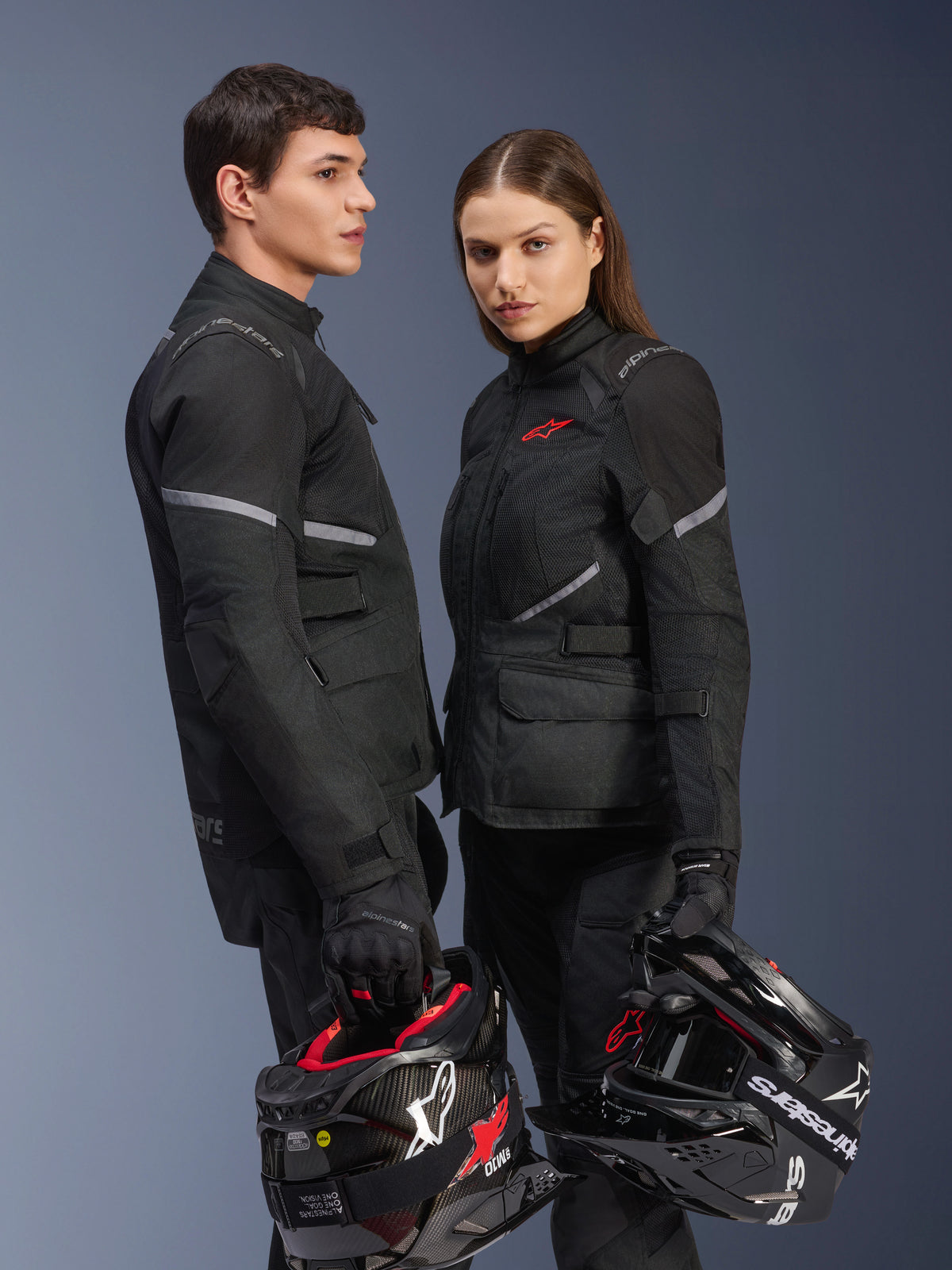 Pilotes homme et femme en équipement moto Alpinestars, présentant la veste Stella Andes Air Drystar® noire, pantalon en textile et gants assortis, tenant des casques noir et blanc, tenue de touring aventure coordonnée, photo en studio