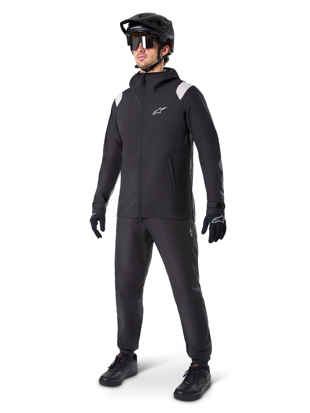 Veste de pluie Alpinestars A-Dura, veste de pluie noire pour VTT avec des détails blancs sur les épaules, portée par un cycliste avec casque de VTT noir, lunettes de soleil, gants et pantalon assortis, tenue de cyclisme technique tout-temps