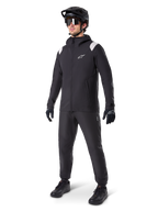 Veste de pluie Alpinestars A-Dura, veste de pluie noire pour VTT avec des détails blancs sur les épaules, portée par un cycliste avec casque de VTT noir, lunettes de soleil, gants et pantalon assortis, tenue de cyclisme technique tout-temps
