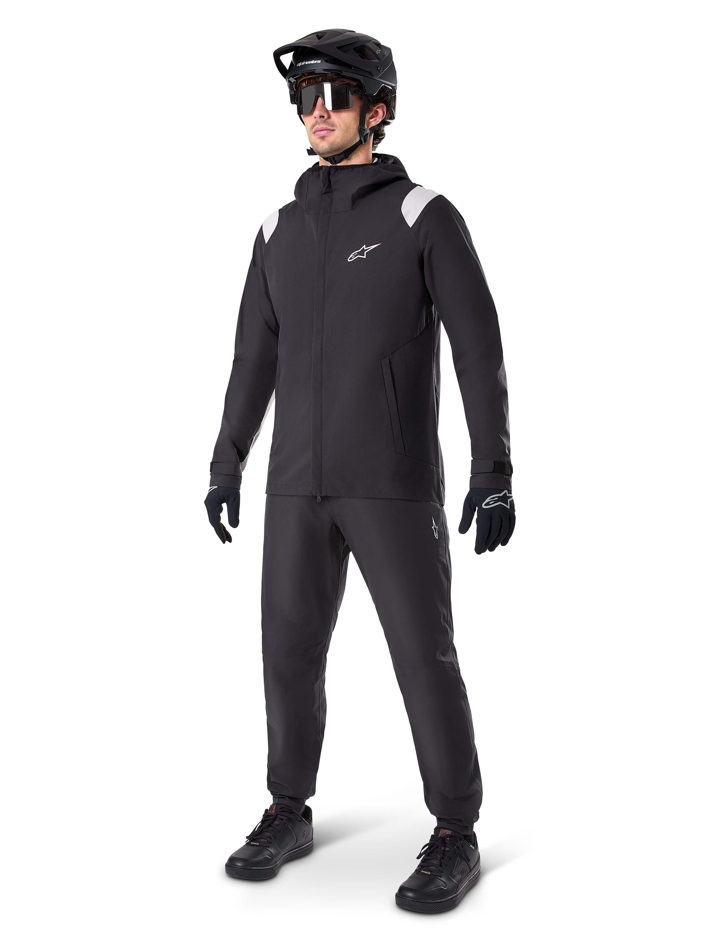 Veste de pluie Alpinestars A-Dura, veste de pluie noire pour VTT avec des détails blancs sur les épaules, portée par un cycliste avec casque de VTT noir, lunettes de soleil, gants et pantalon assortis, tenue de cyclisme technique tout-temps
