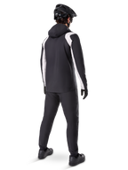 Veste de pluie Alpinestars A-Dura, vue arrière d'un cycliste portant la veste de VTT imperméable noire et blanche, pantalon de cyclisme noir assorti, casque et gants noirs, équipement technique pour le VTT par conditions humides