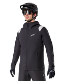 Veste de pluie Alpinestars A-Dura, veste de pluie pour VTT, noire avec accents gris clair sur les épaules, portée par un cycliste avec un casque de VTT noir et des lunettes de soleil, logo blanc sur la poitrine, construction imperméable, coordonnée avec un pantalon et des gants noirs pour le cyclisme tout-terrain