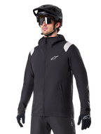 Veste de pluie Alpinestars A-Dura, veste de pluie pour VTT, noire avec accents gris clair sur les épaules, portée par un cycliste avec un casque de VTT noir et des lunettes de soleil, logo blanc sur la poitrine, construction imperméable, coordonnée avec un pantalon et des gants noirs pour le cyclisme tout-terrain
