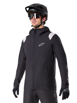 Alpinestars A-Dura Regenjacke, Mountainbike-Regenjacke, Schwarz mit hellgrauen Schulterakzenten, getragen von einem Fahrer mit schwarzem MTB-Helm und Sonnenbrille, weißes Logo auf der Brust, wasserdichte Konstruktion, kombiniert mit schwarzer Hose und Handschuhen für den Offroad-Radsport