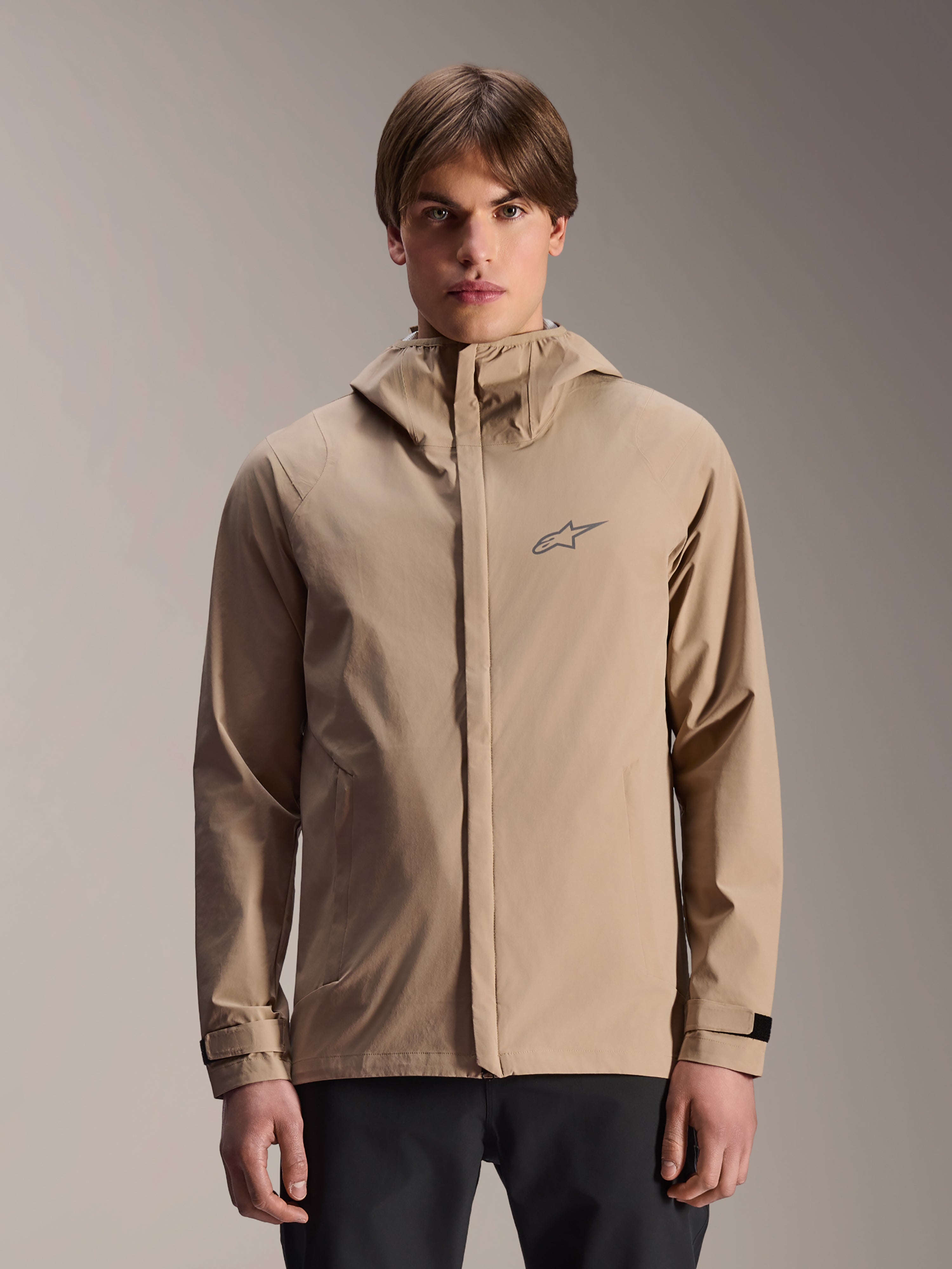 Veste de pluie Alpinestars A-Dura, veste de pluie de VTT à capuche marron clair, portée par un mannequin, dotée d'un petit logo sur la poitrine, de poignets réglables et d'une fermeture éclair frontale dissimulée, équipement de cyclisme imperméable