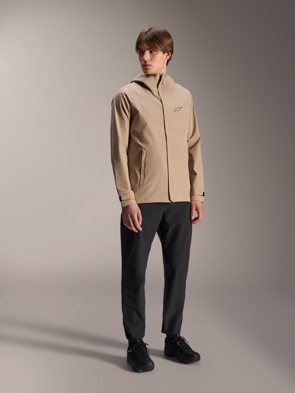 Männliches Model in der Alpinestars A-Dura Regenjacke in Hellbraun, kombiniert mit schwarzer Hose und Schuhen, minimalistische Outdoor-Mountainbike-Regenjacke mit Kapuzendesign und kleinem Logo auf der Brust.