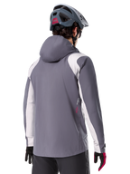 Veste de pluie Alpinestars A-Dura, veste de VTT, gris titane foncé, vue de dos avec empiècements gris clair sur les épaules et les bras, détail du logo rouge sur le côté, portée avec un casque de VTT gris et des gants noirs et rouges, vêtement technique de trail