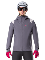 Veste de pluie Alpinestars A-Dura, veste de pluie pour VTT, gris titanium foncé, modèle à capuche avec logo Alpinestars rose et empiècements gris clair aux épaules, portée avec un casque de VTT blanc et des gants rose vif, équipement imperméable pour le cyclisme tout-terrain